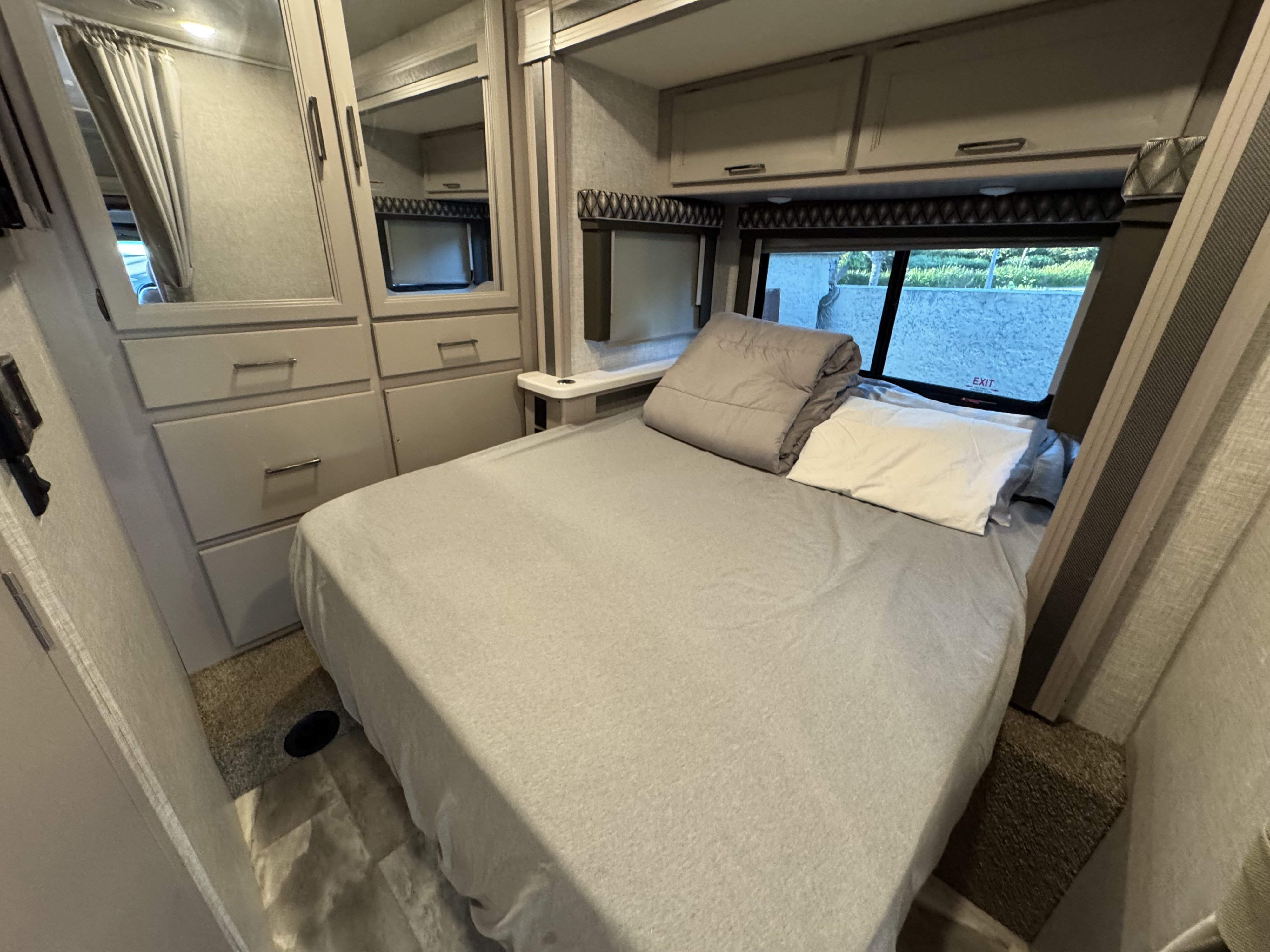 This RV rental features a comfortable and modern bedroom with a bed, mirrored wardrobes, overhead cabinets, and a window.. Thor Quantum 2022