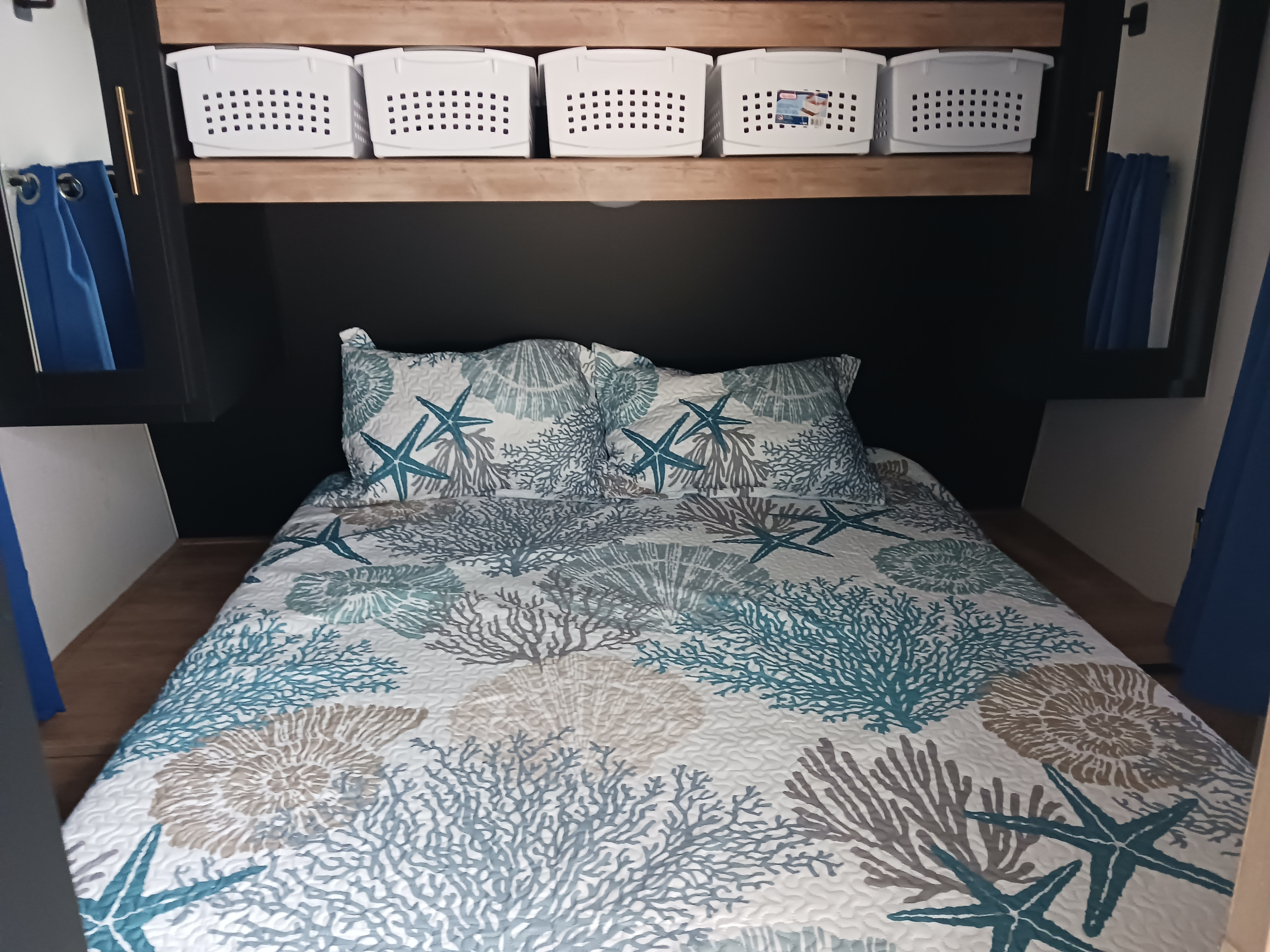 Cozy RV rental bedroom with a coastal-themed bedspread, pillows, and organized overhead storage baskets.. Keystone RV Springdale 2023