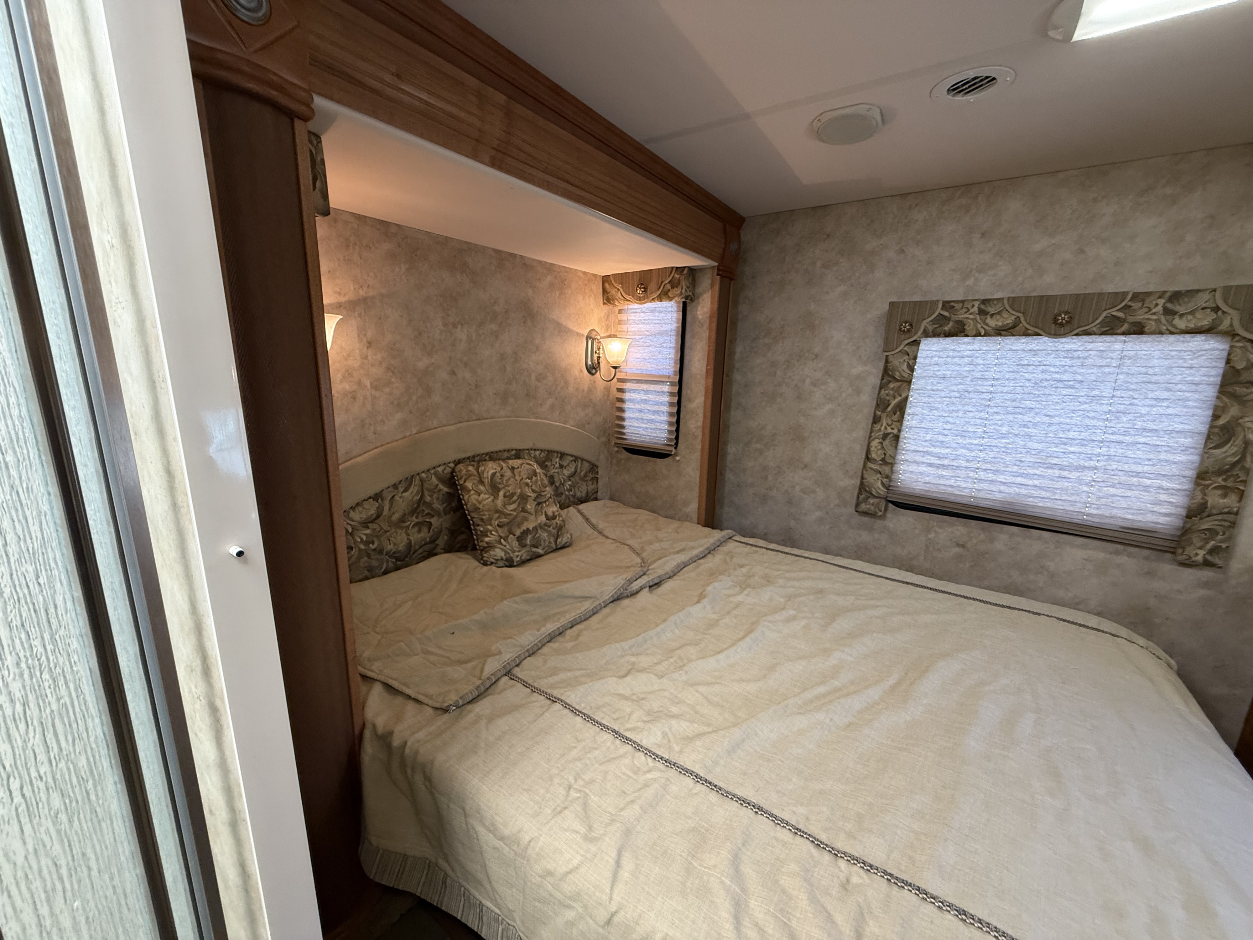 RV rental's inviting bedroom: soft bedding, decorative headboard, wall sconces, and two windows with blinds.. Gulf Stream Independence 2010