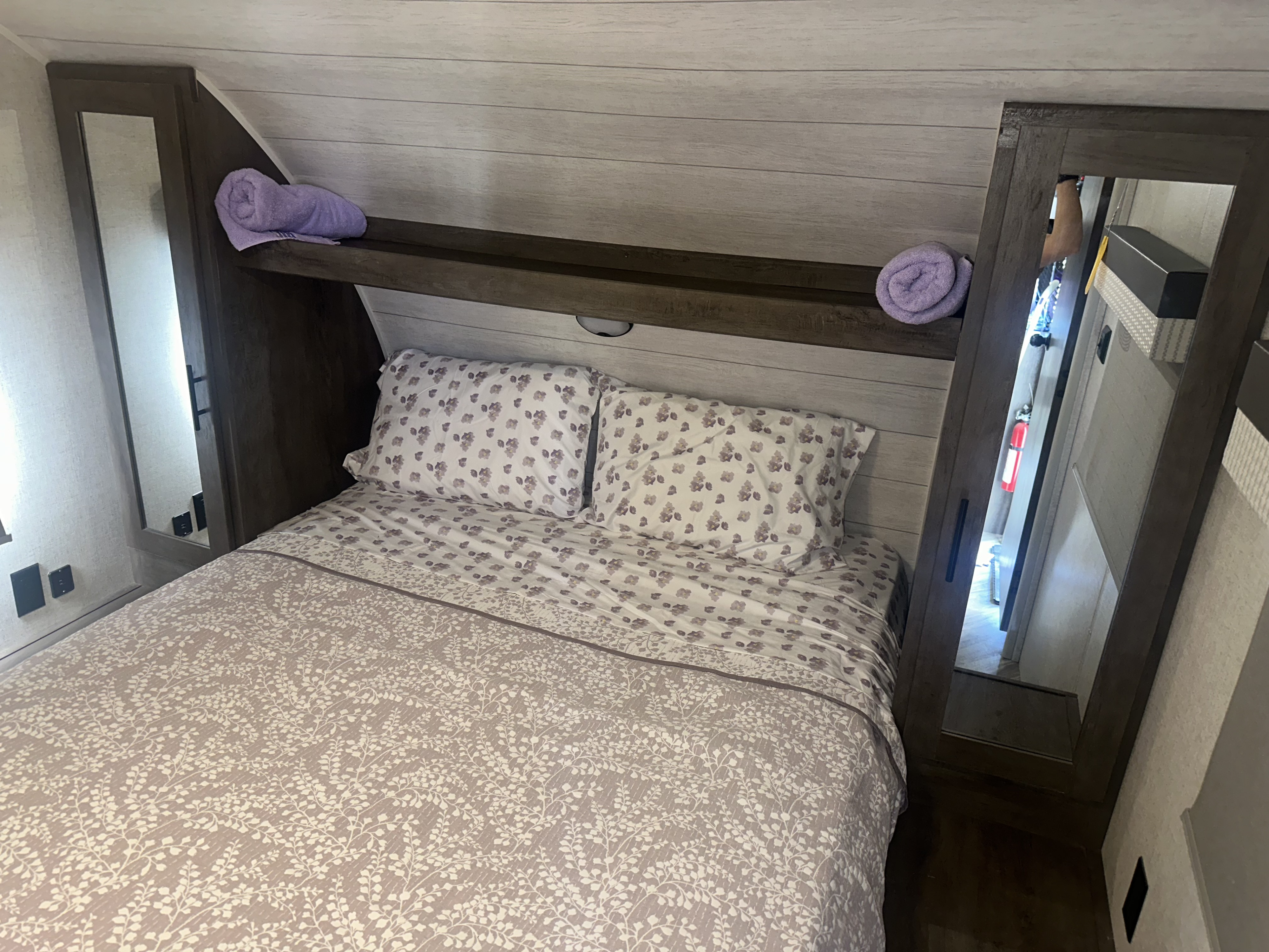 This RV rental bedroom offers a cozy bed with floral patterns, mirrored storage, and a shelf with purple towels, perfect for your trip.. Forest River Wildwood X-Lite 2022
