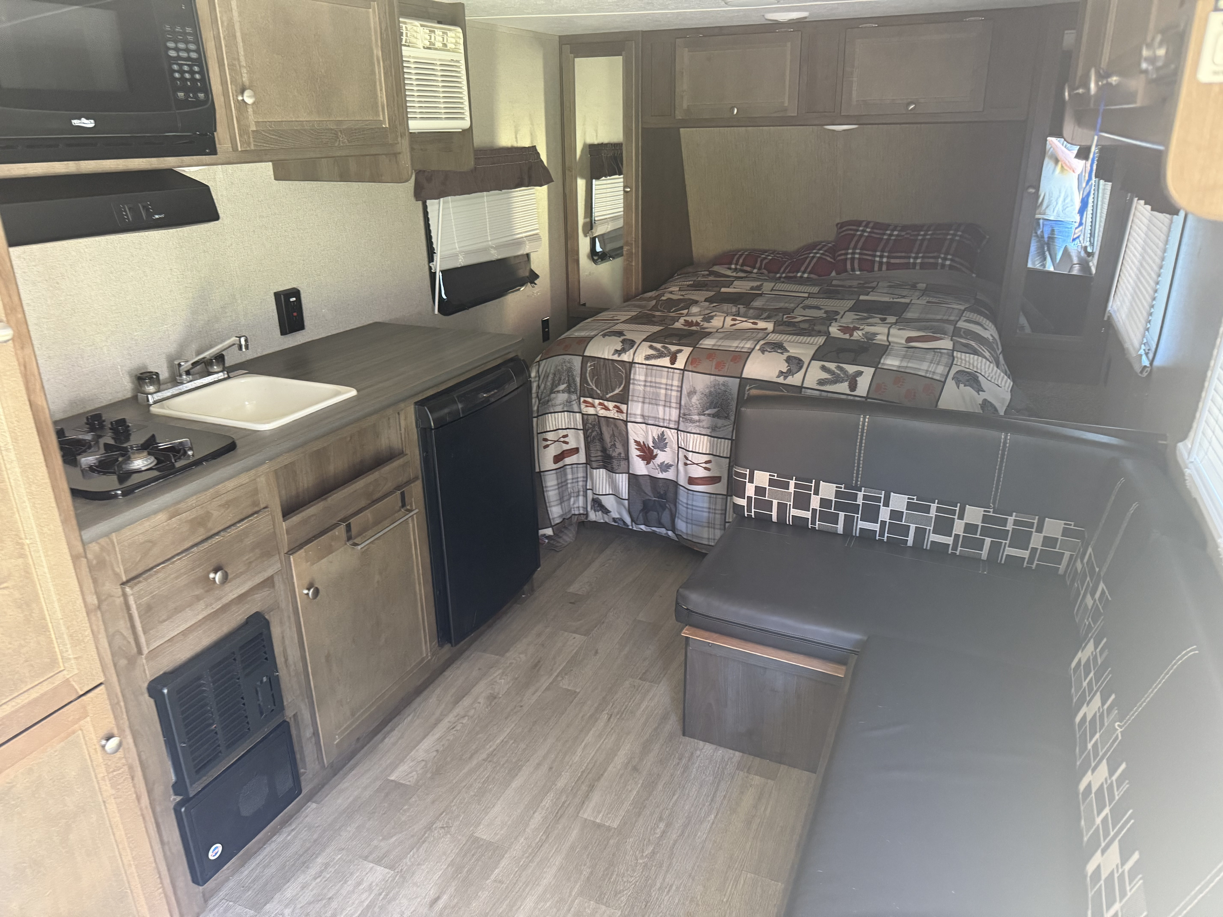 RV rental interior: kitchen with appliances on left, bed with plaid comforter in center, dark seating area on right.. Coleman Lantern 2018