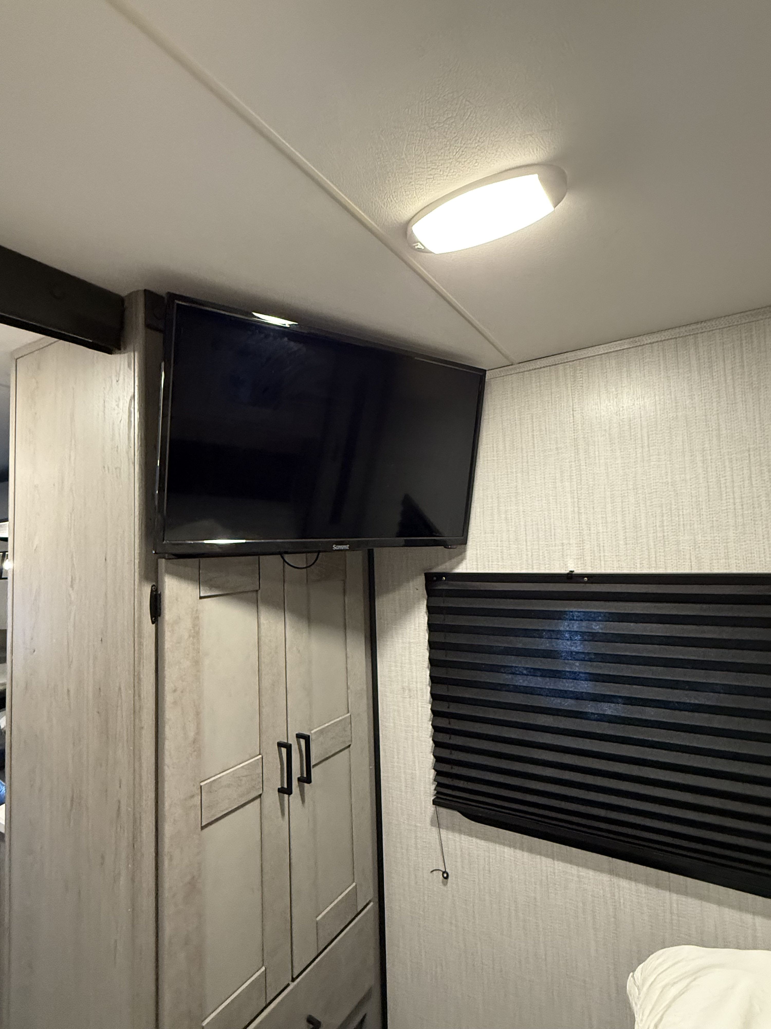 RV rental bedroom featuring a wall-mounted TV, light cabinet, window blind, and bright ceiling light.. Heartland RVs Mallard 2021