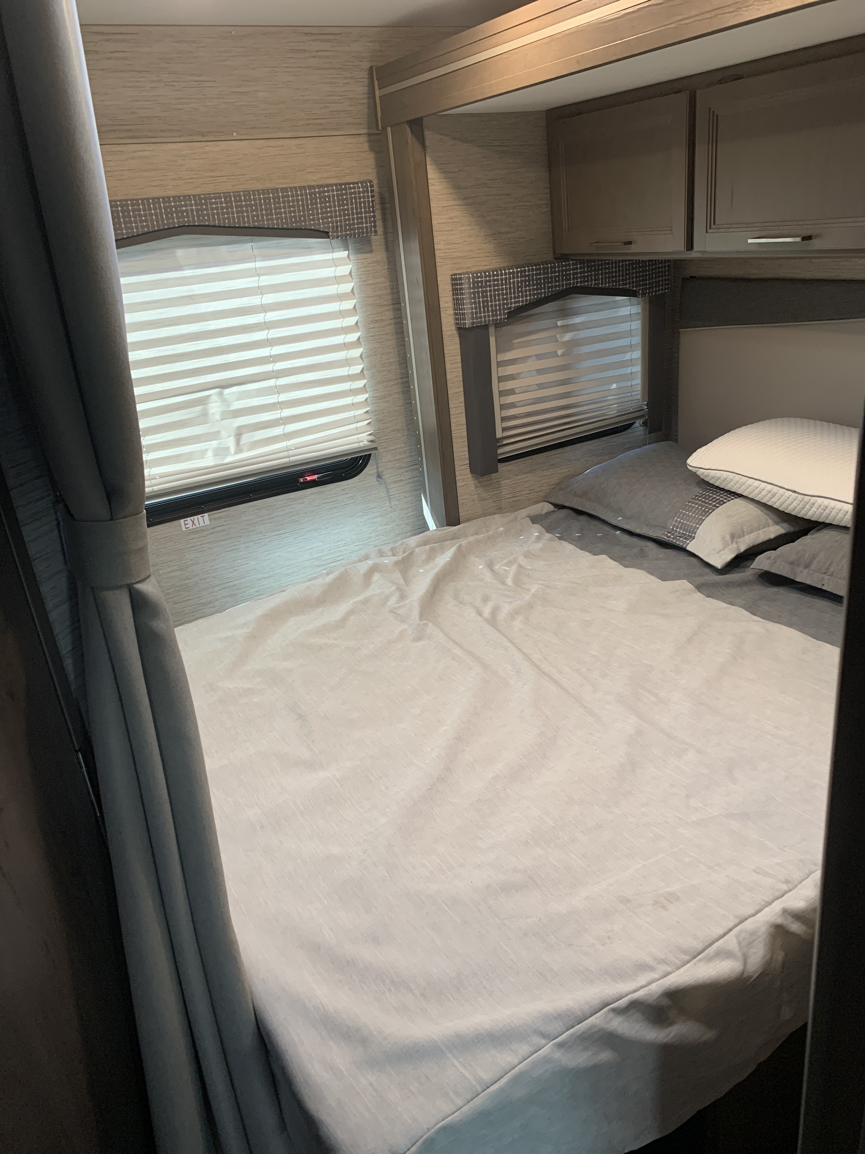Interior of an RV rental bedroom: A comfortable bed with light bedding, windows with blinds, and overhead cabinets.. Thor Chateau 2022