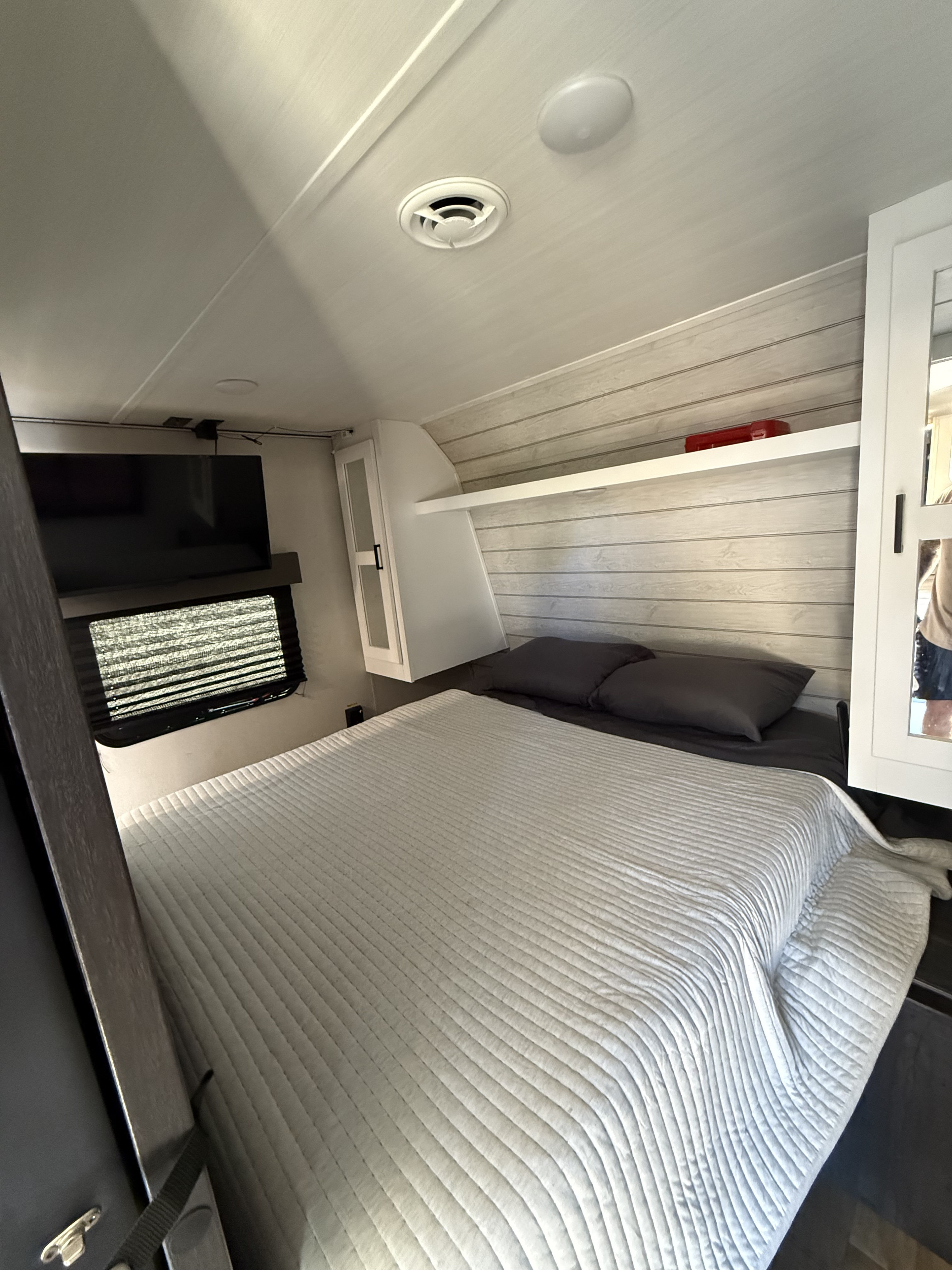 Inside an RV rental, a bedroom with a cozy bed, wall-mounted TV, and overhead cabinets.. Keystone RV Hideout 2023
