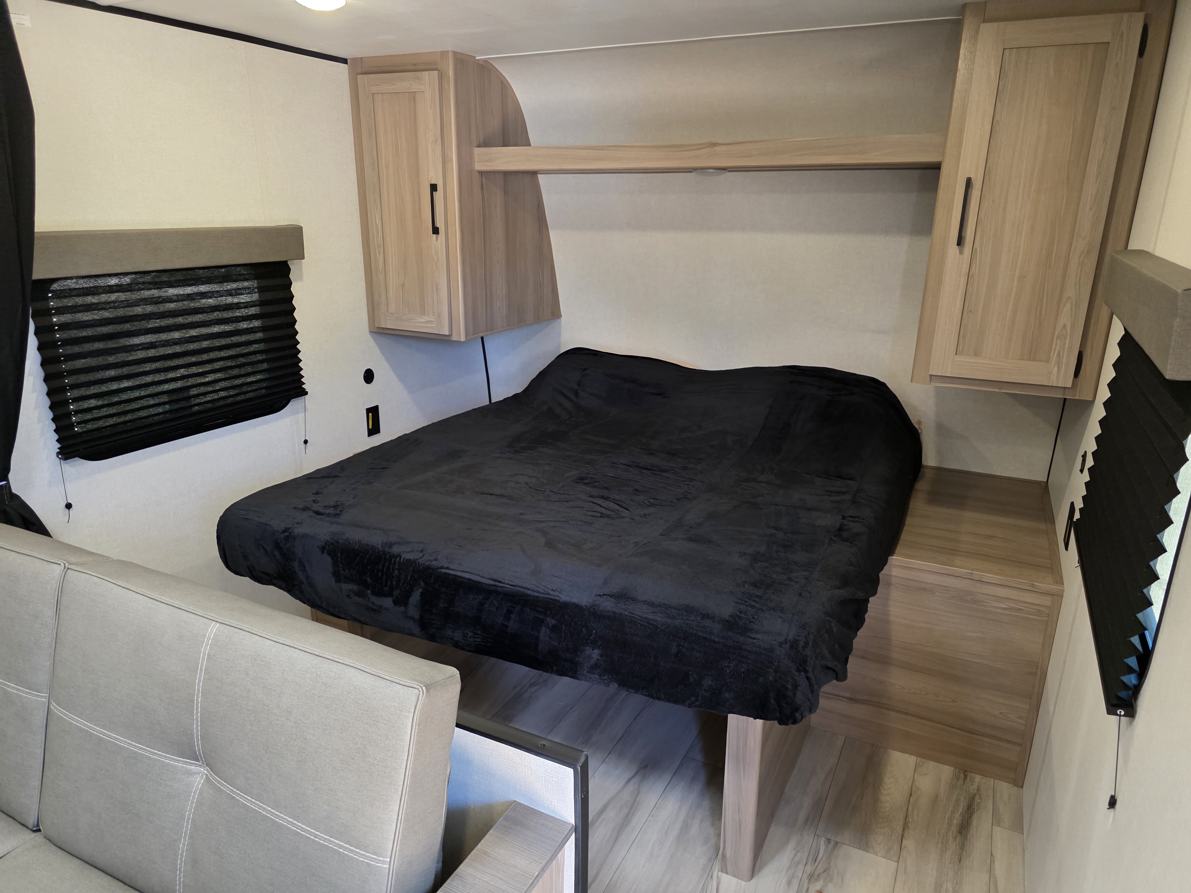 This RV rental features a cozy bedroom with a black blanket on the bed, overhead cabinets, a window, and a sofa.. Keystone Coleman 2025