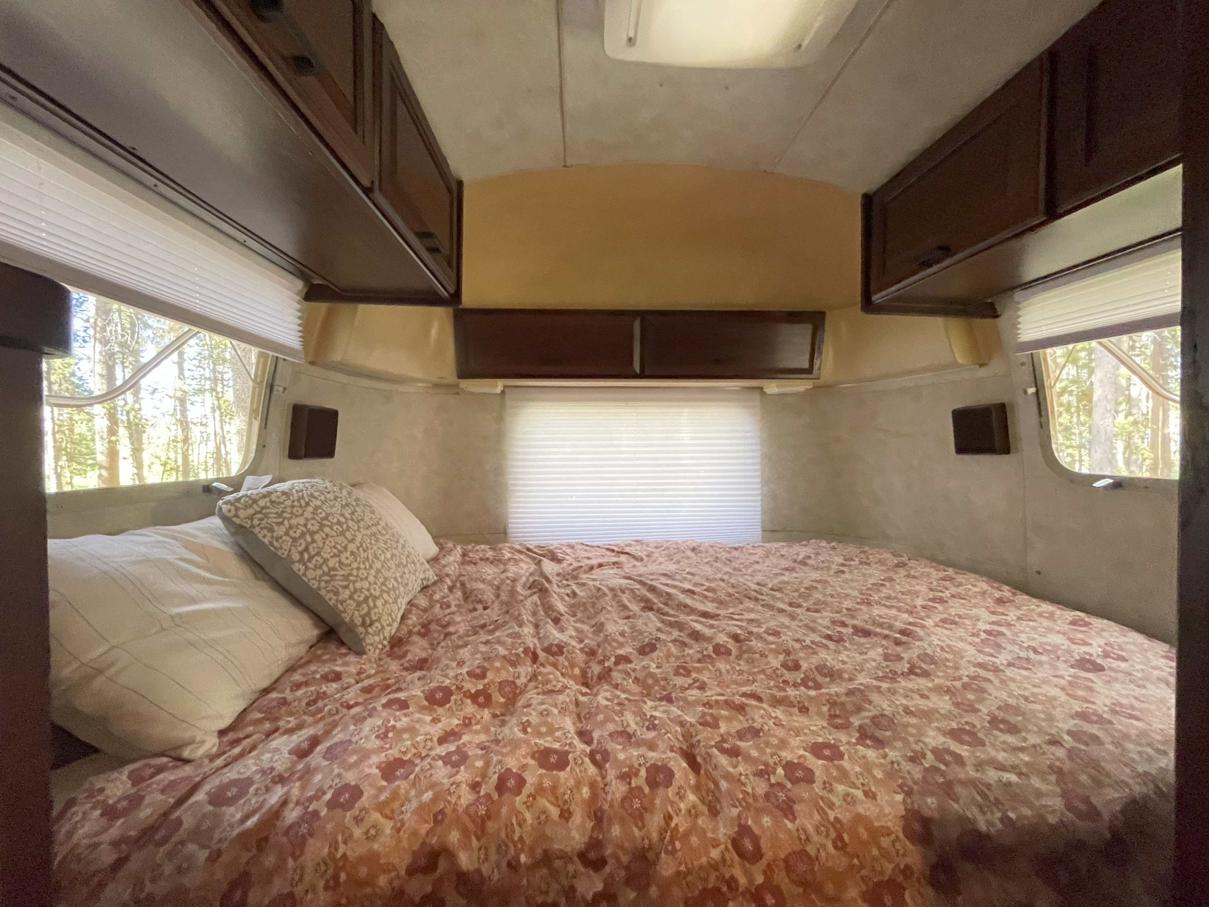 Cozy RV rental bedroom with a floral comforter, overhead cabinets, and large windows revealing a peaceful forest view.. Airstream Excella 1990