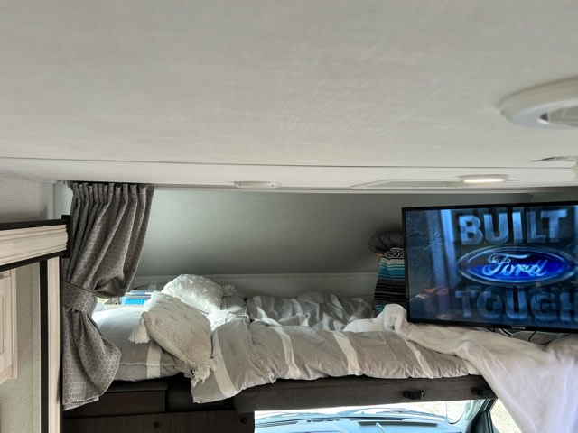 Inside an RV rental: a comfortable cab-over bed area with bedding, pillows, and a TV showing a Ford ad.. Forester LE 2021