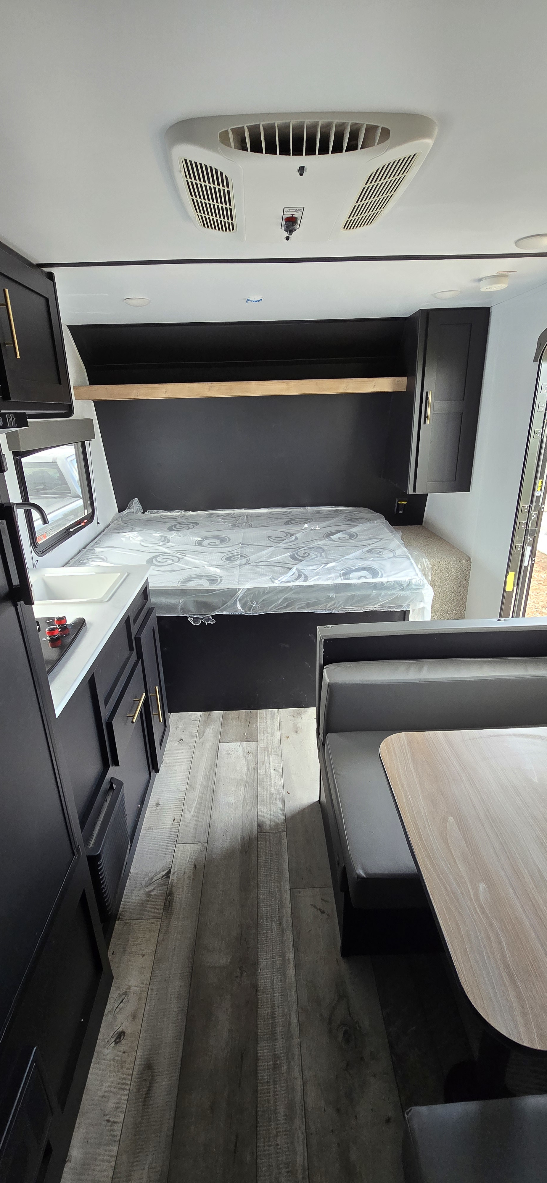 RV rental interior: plastic-wrapped bed, kitchenette, dinette, and overhead AC unit. Modern black and wood tones.. Keystone RV Springdale 2022