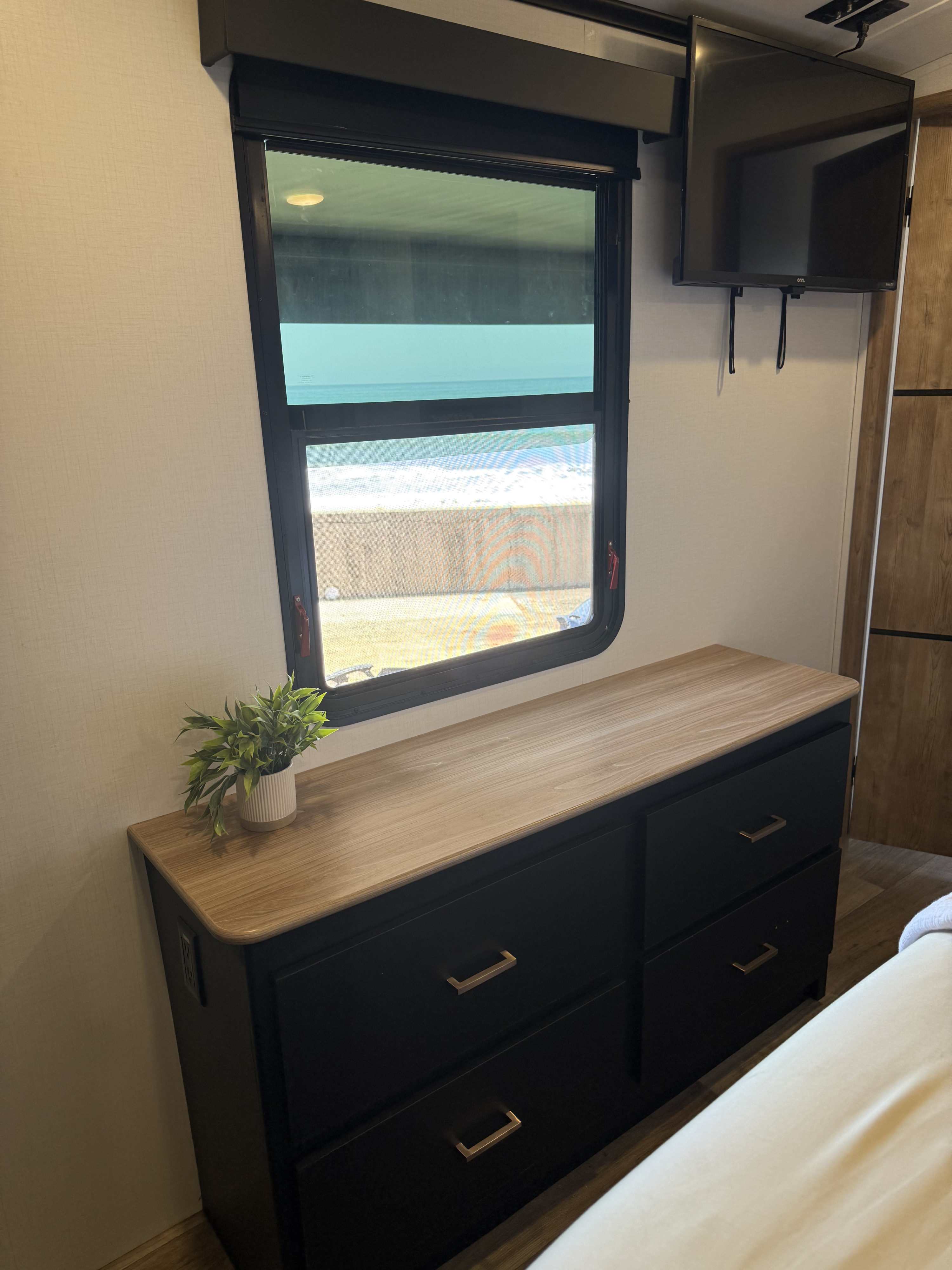 Inside an RV rental: a cozy bedroom setup with a dresser, wall-mounted TV, and a scenic window view of the ocean.. Keystone RV Outback 2023