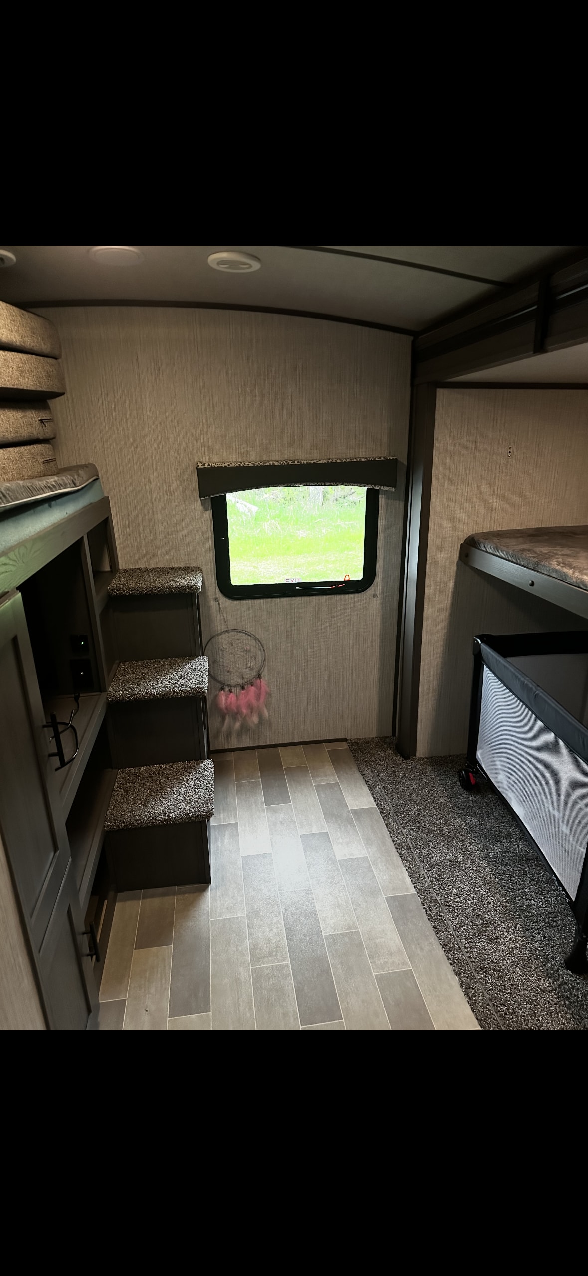 Interior of an RV rental bunk room: multiple beds with steps, a window showing greenery, and a portable baby crib on carpet.. Cruiser RV MPG Ultra Lite 2021
