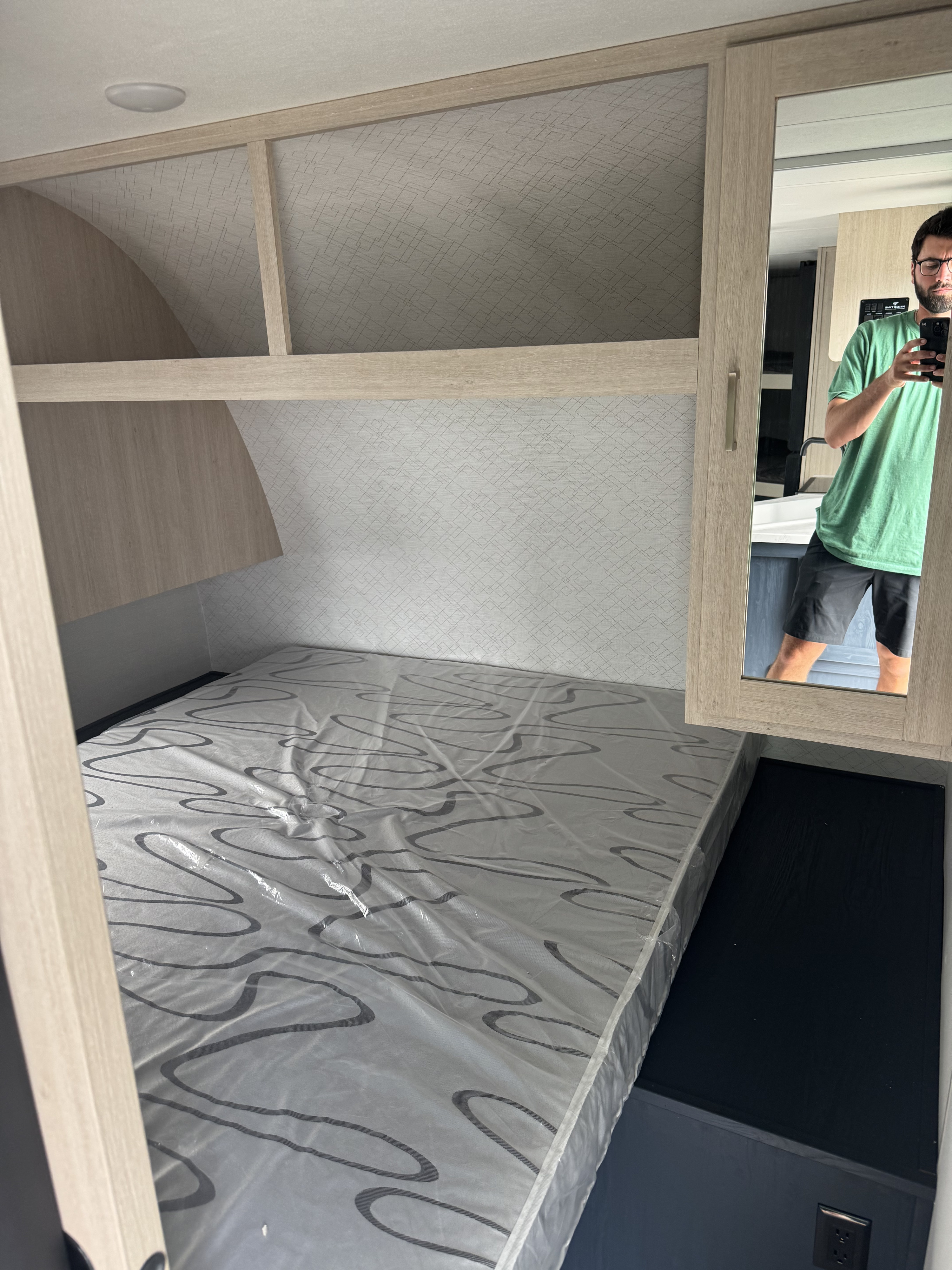 Compact RV rental sleeping area featuring a plastic-wrapped mattress, overhead storage, and mirror reflecting the owner and sink.. Prime Time Manufacturing Avenger LE 24BHSLE 2025