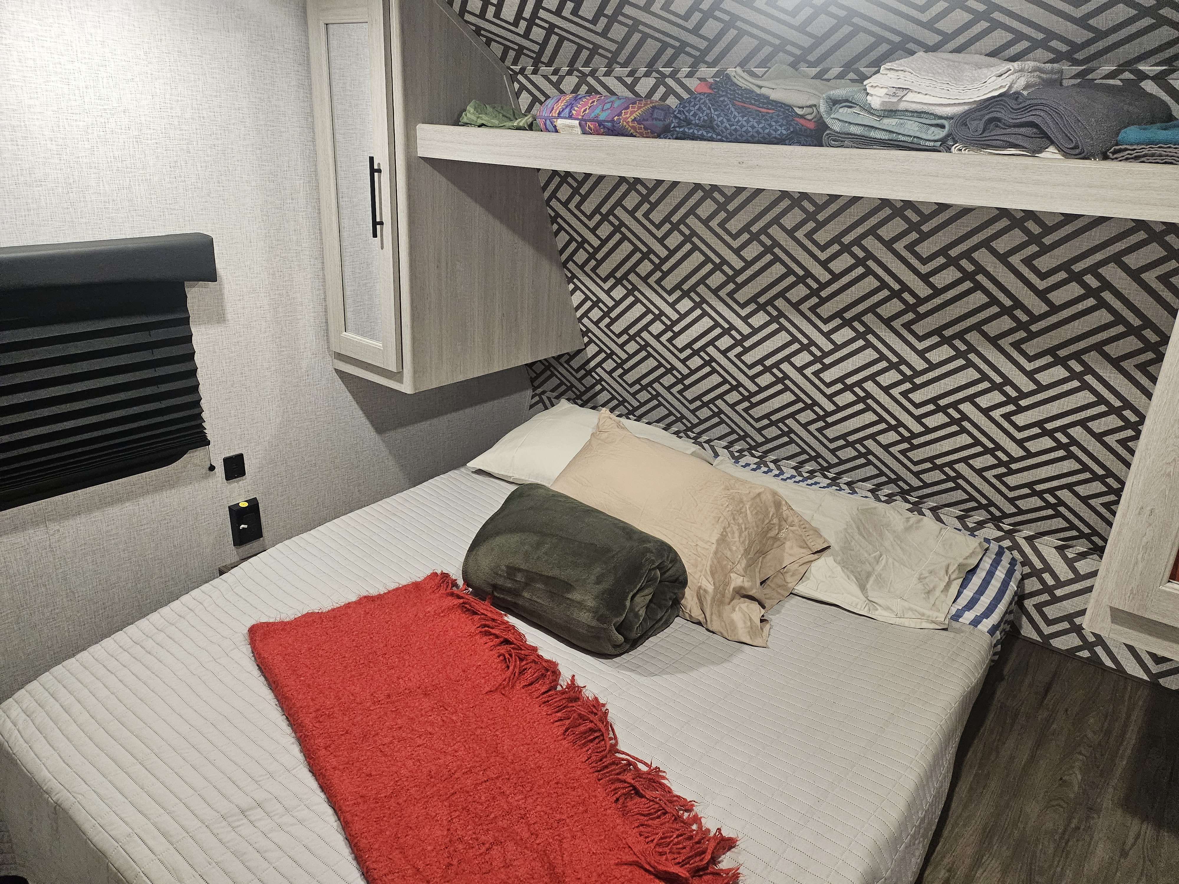 Cozy RV rental bedroom featuring a bed with colorful blankets, a patterned accent wall, and overhead storage with neatly folded items.. Crossroads RV ZR270BH 2024