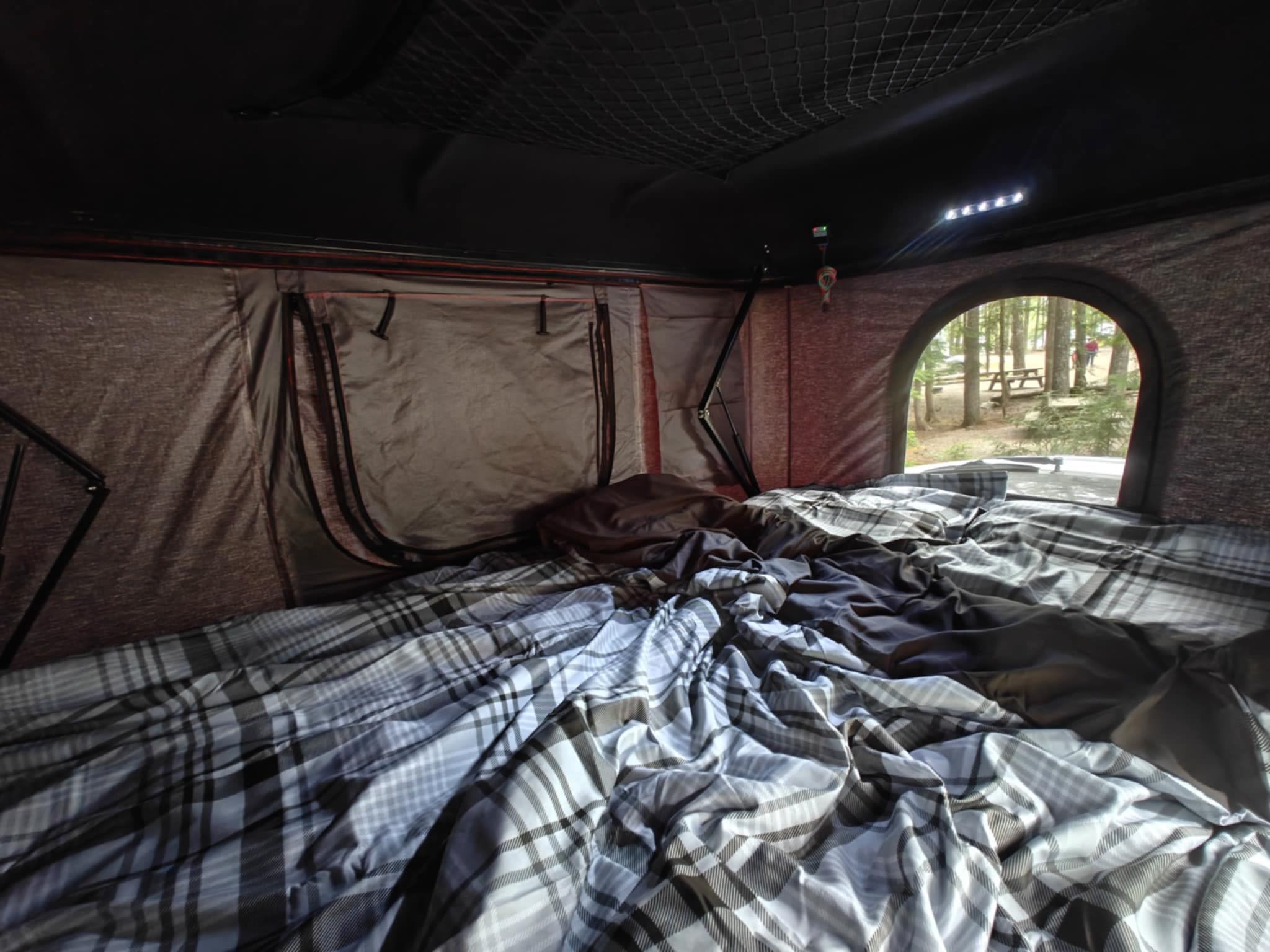 Enjoy the cozy interior of this RV rental's rooftop tent! Plaid bedding, overhead net, and a lovely forest campsite view.. Dodge Grand Caravan 2011