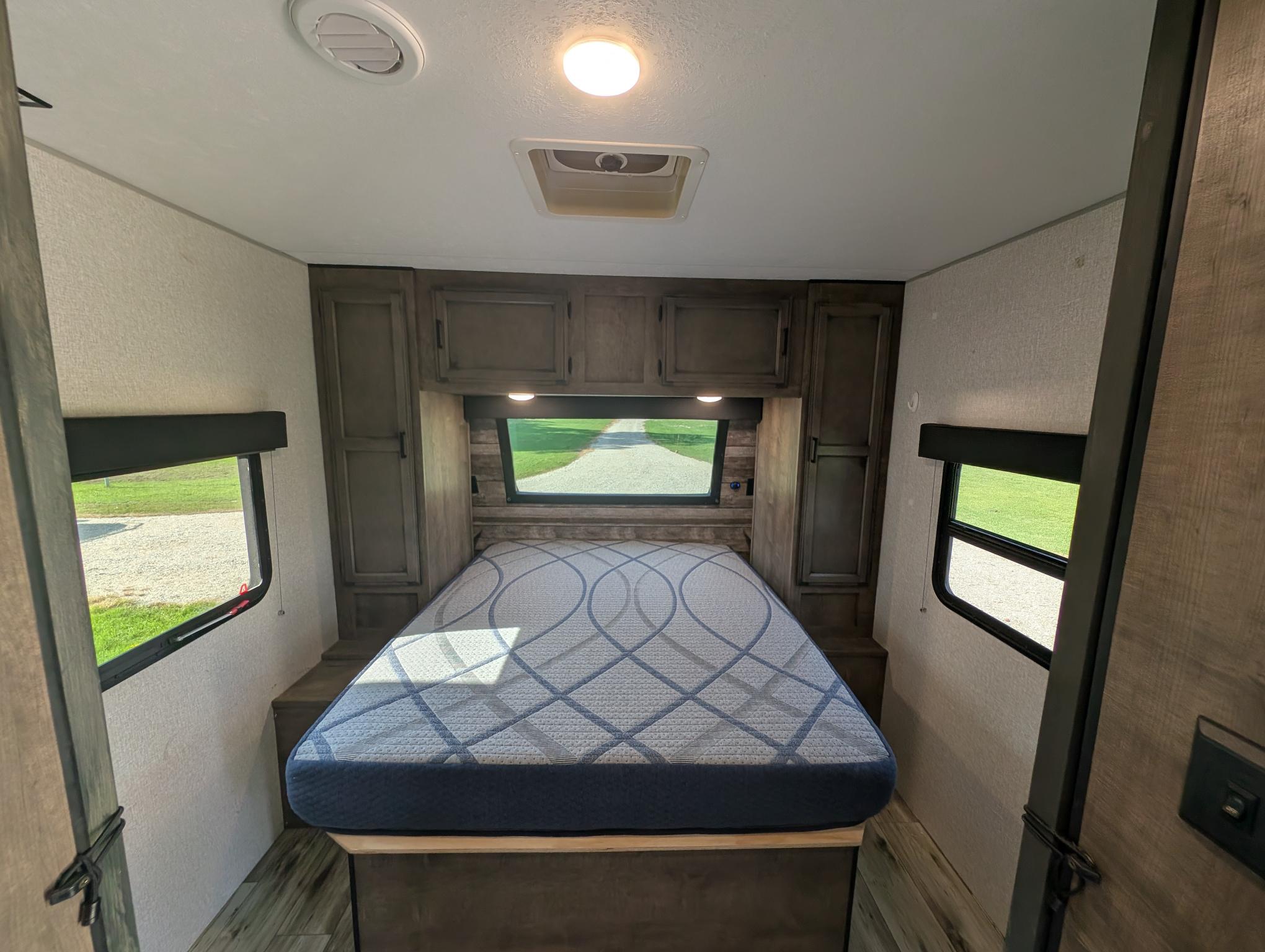 Cozy RV rental bedroom featuring a large bed with patterned mattress, overhead storage, and side windows showcasing outdoor views.. Venture 332VBH 2022