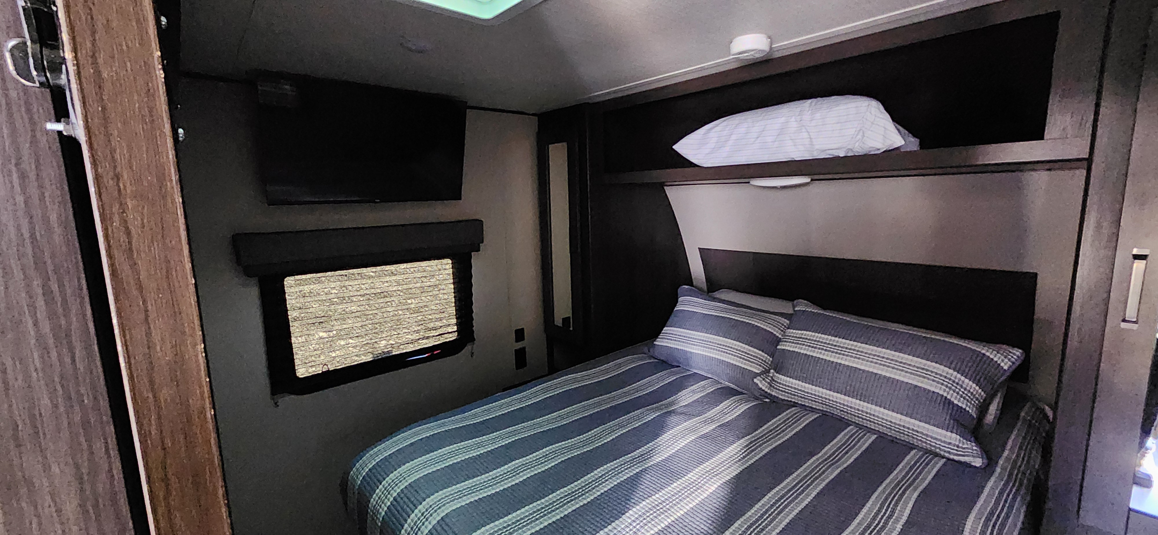 Cozy RV rental bedroom interior, featuring a bed with striped linens, TV, and window with blinds.. Grand Design Transcend Xplor 2022