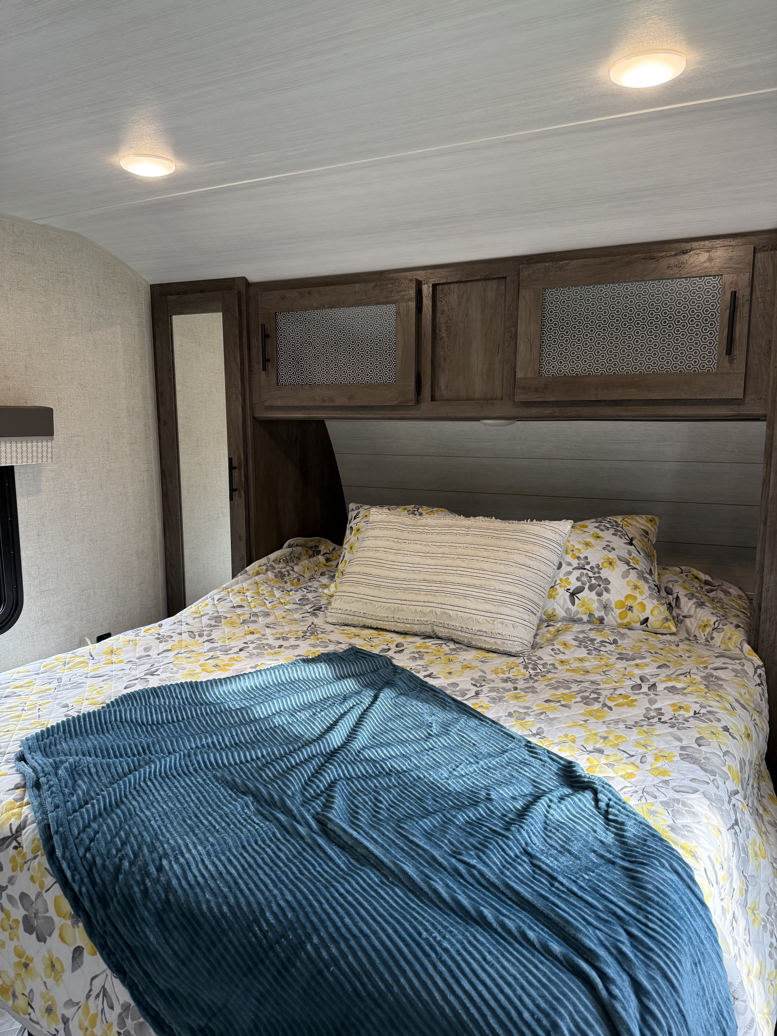 Bright RV rental bedroom interior featuring a comfortable bed with floral bedding, a blue blanket, and built-in wood cabinets.. Forest River Wildwood 2021