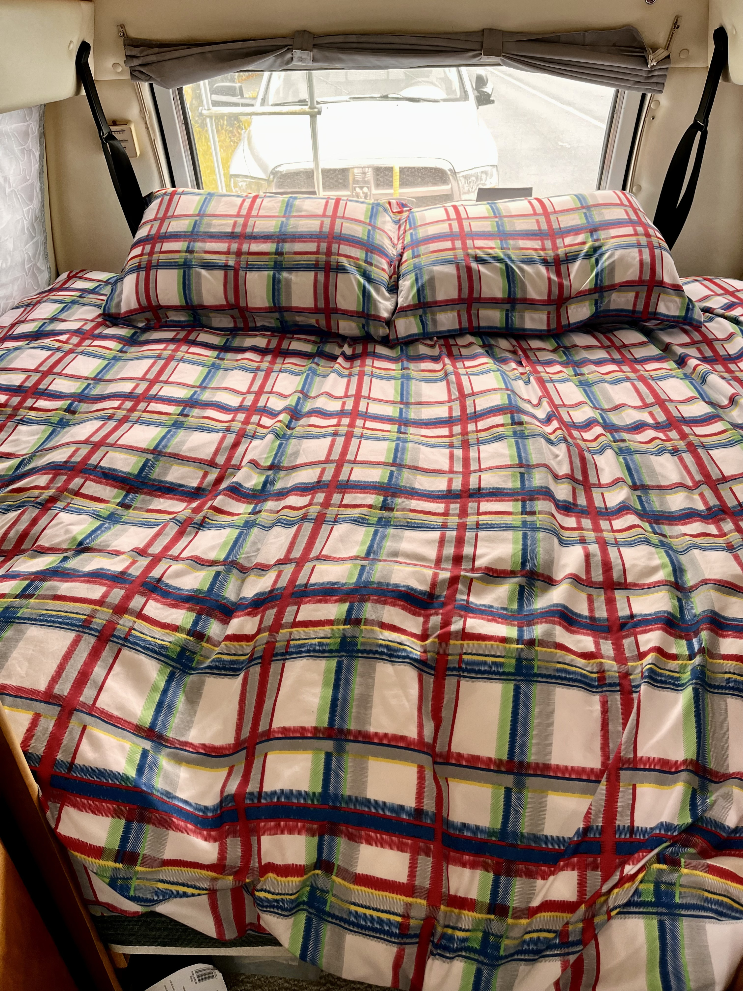Cozy RV rental interior features vibrant plaid bedding, with a view of a parked truck through the rear window.. Dodge Leisure Travel Wide Body 2002