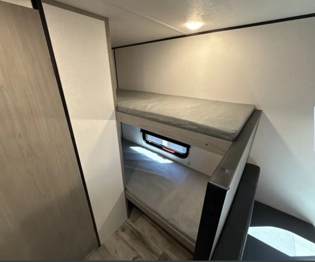 Comfortable bunk beds in an RV rental interior, showing compact sleeping for two. Light walls, small window on bottom bunk.. Coleman Lantern 17b 2024