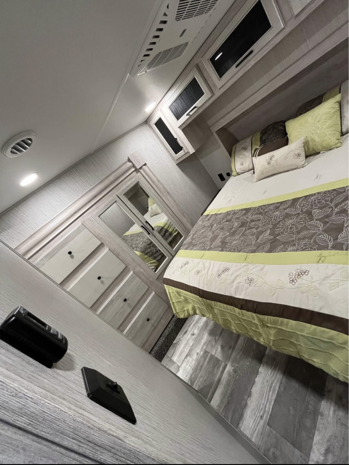 Modern RV rental bedroom, showing a bed with green/brown bedding, overhead cabinets, AC, a mirrored wardrobe, and gray wood-look floors.. Forest River Sierra 2023
