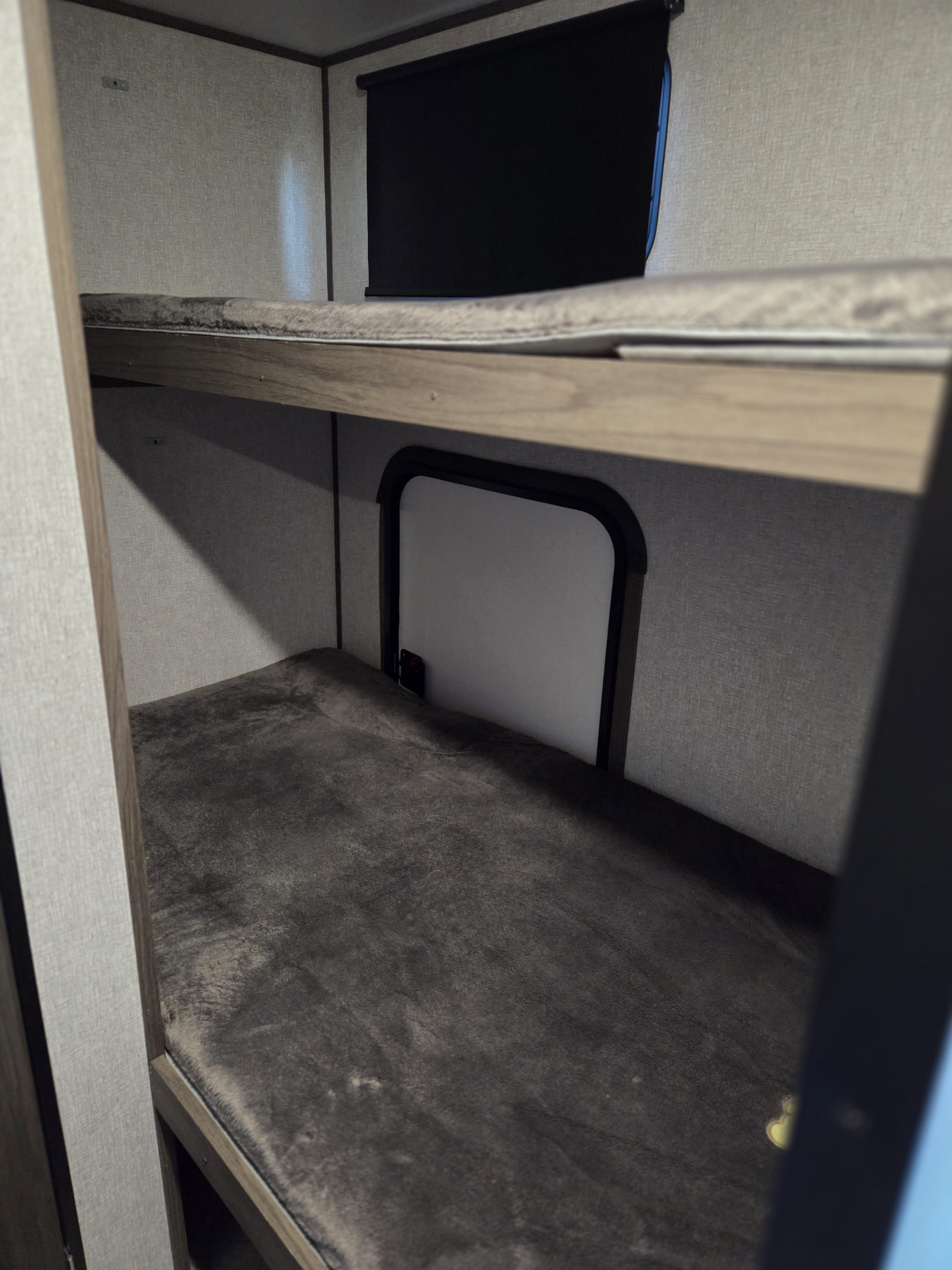 Inside an RV rental, cozy bunk beds with dark grey bedding. An upper window with a black shade and a lower access hatch are visible.. Dutchmen Kodiak Cub 2021