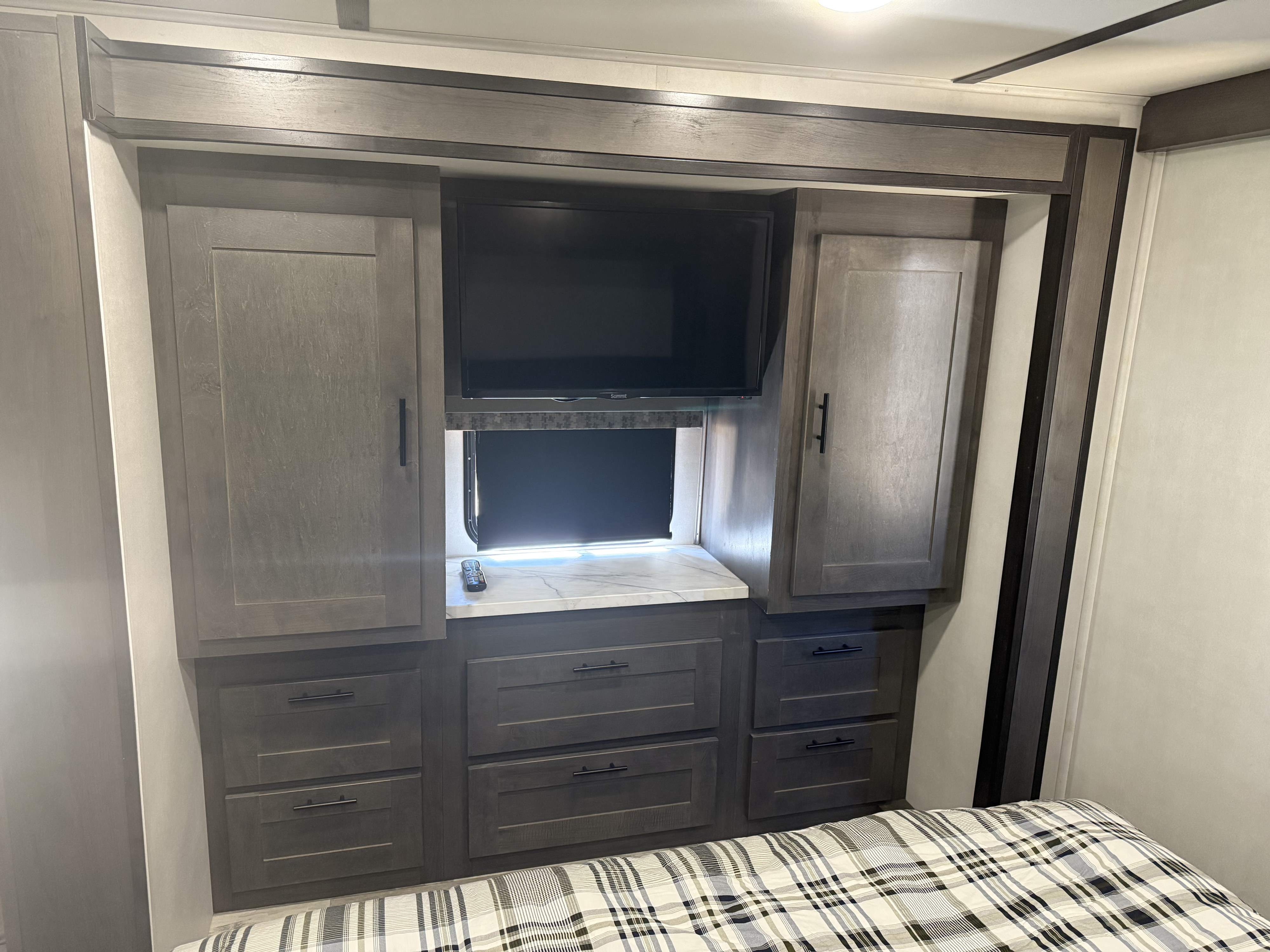 Modern RV rental bedroom with built-in TV, surrounding cabinets, and drawers, visible above a plaid bed.. FR3 FR3 Motorhome 2021
