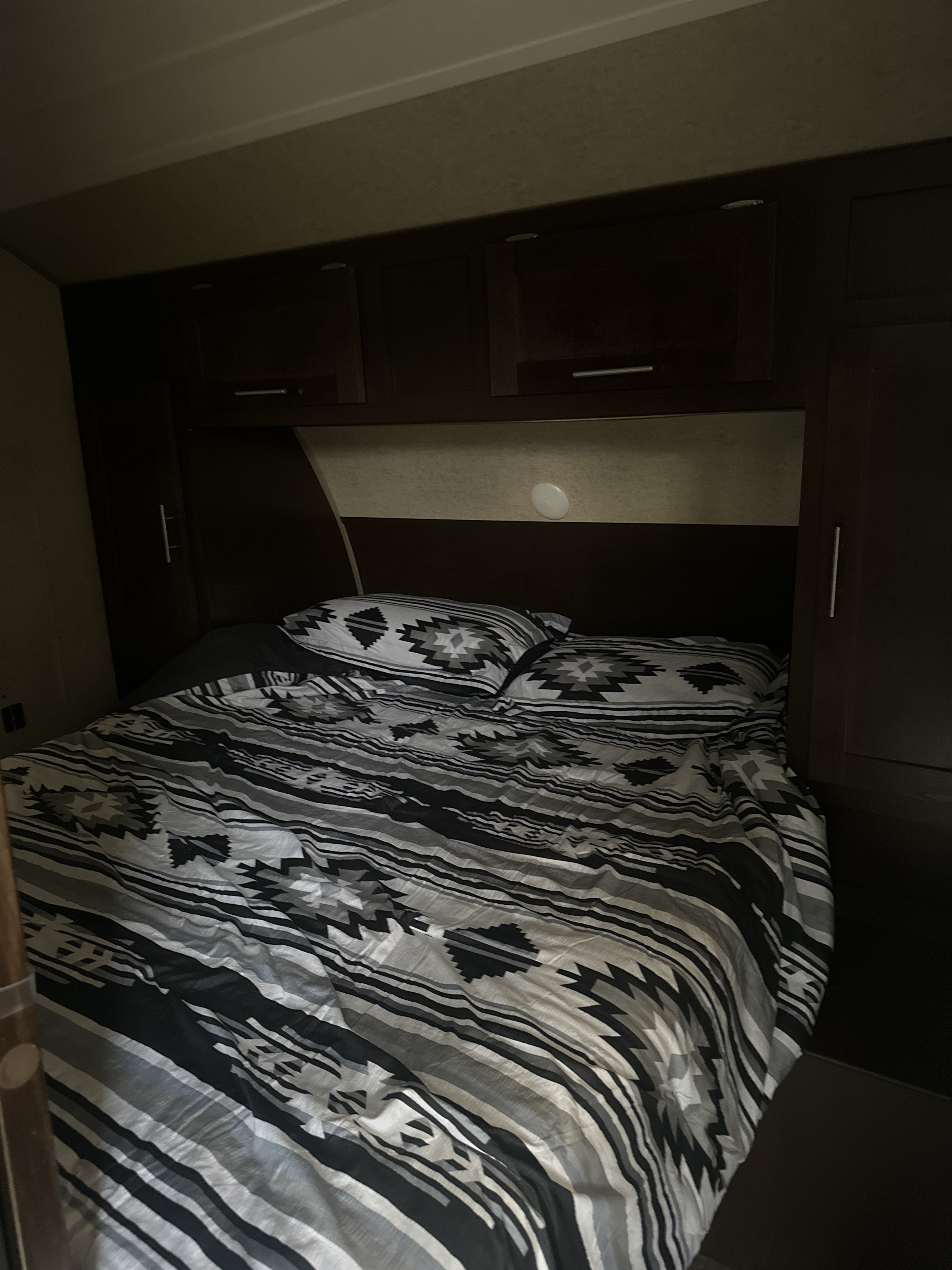 This RV rental bedroom has a bed with patterned black, white, and grey bedding, plus dark wood overhead cabinets.. Forest River Cherokee Grey Wolf 2016