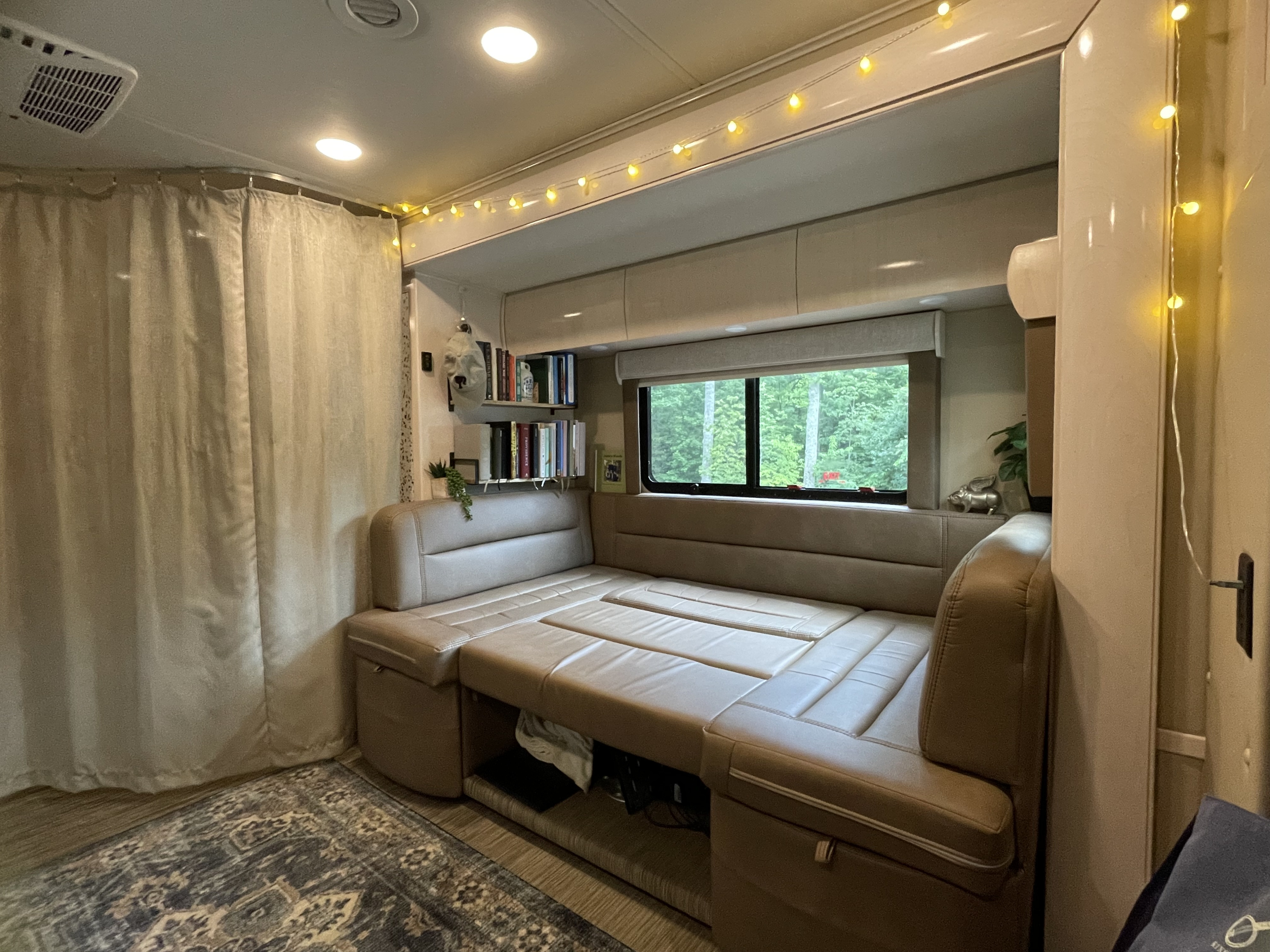 Cozy RV rental interior featuring a convertible dinette bed, window with nature view, bookshelves, and string lights.. Renegade Custom Coaches Vienna 2021