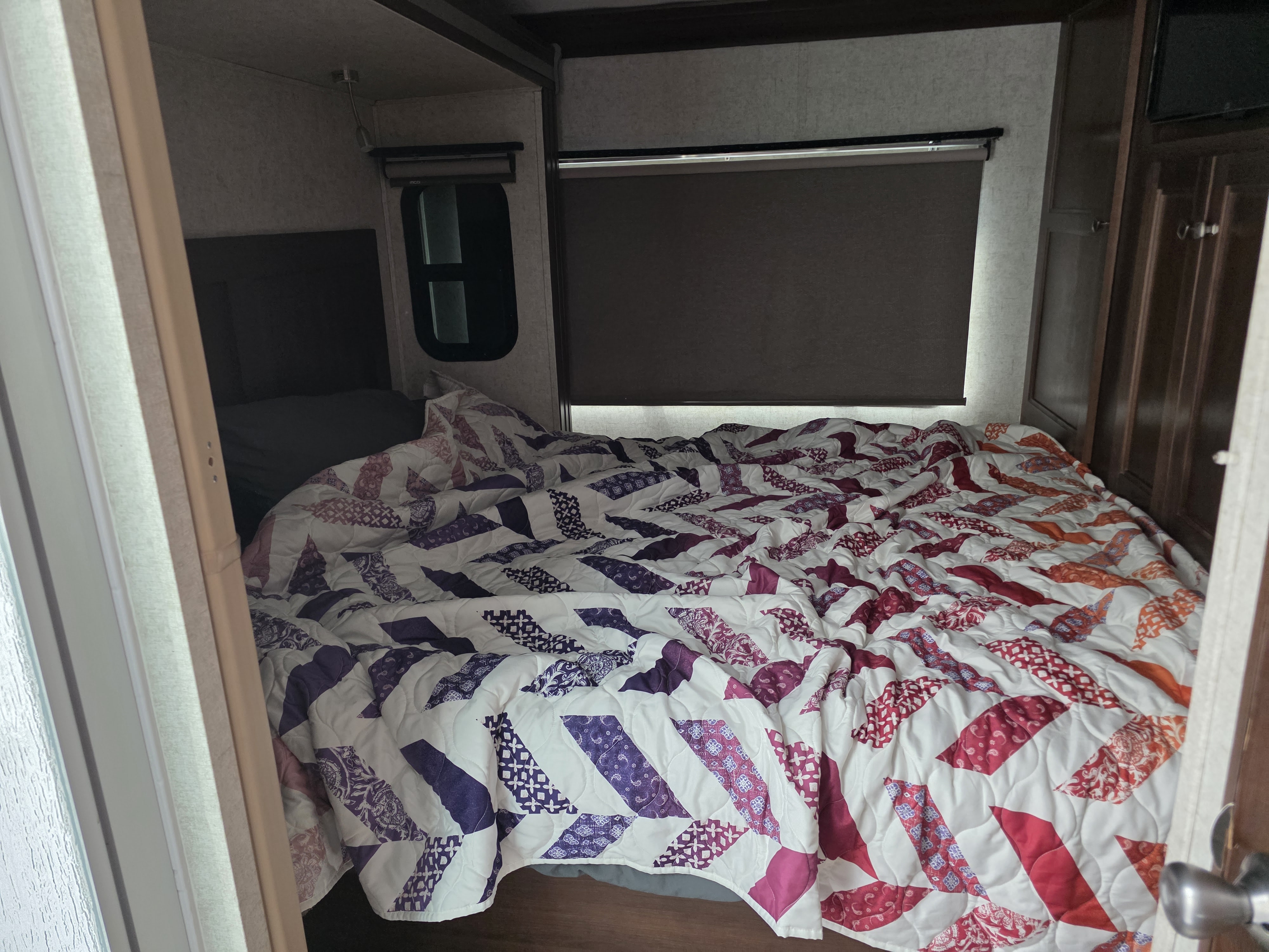 View into an RV rental bedroom showing a bed with a colorful quilt, compact windows, and wood cabinetry.. Forest River Forester 2019
