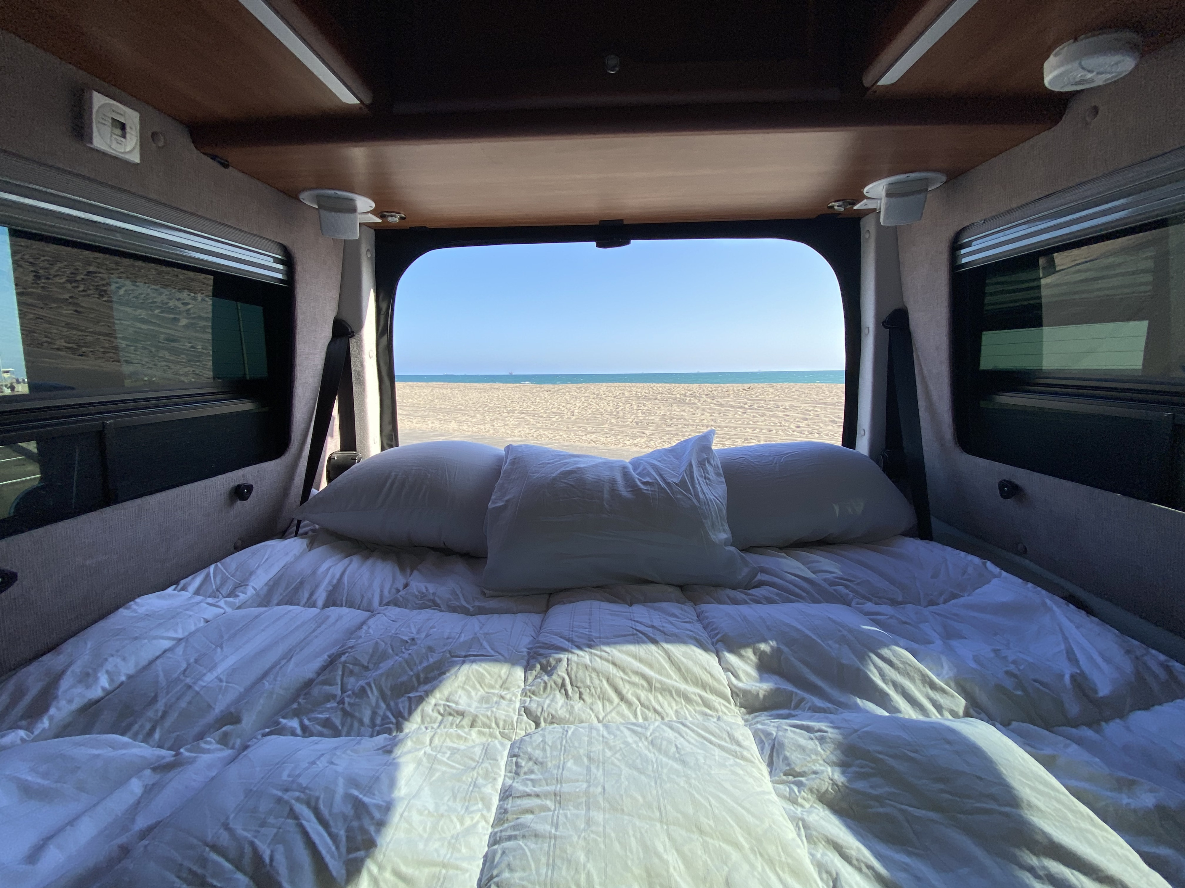 Cozy rv rental bed with white linens and pillows, framed by the open back, reveals a stunning sandy beach & clear ocean view.. Roadtrek Roadtrek Class B 2015