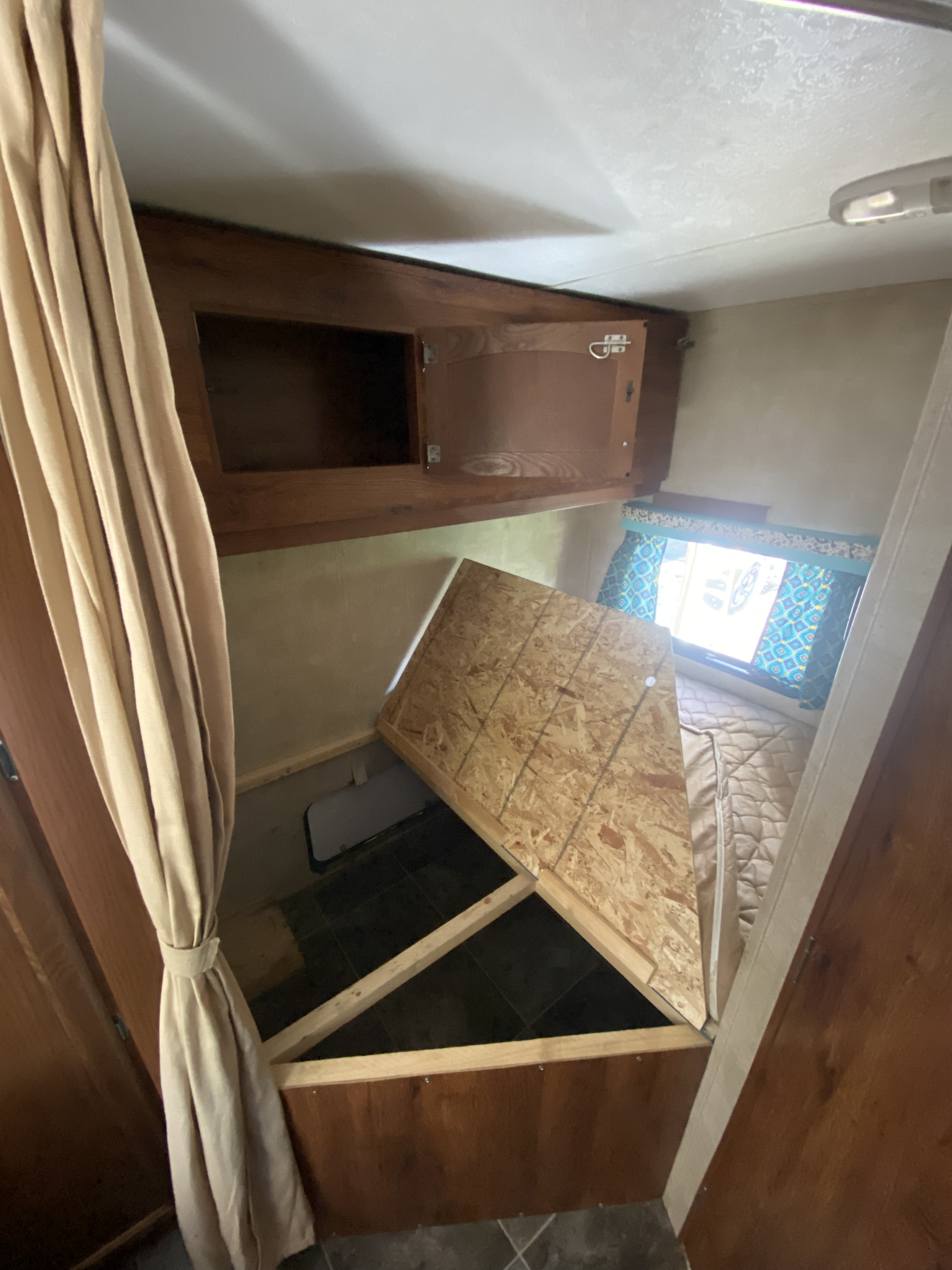 RV rental interior with a lifted bed platform, revealing hidden storage. Features an open cabinet, window, and patterned curtains.. Keystone RV Hideout 2015