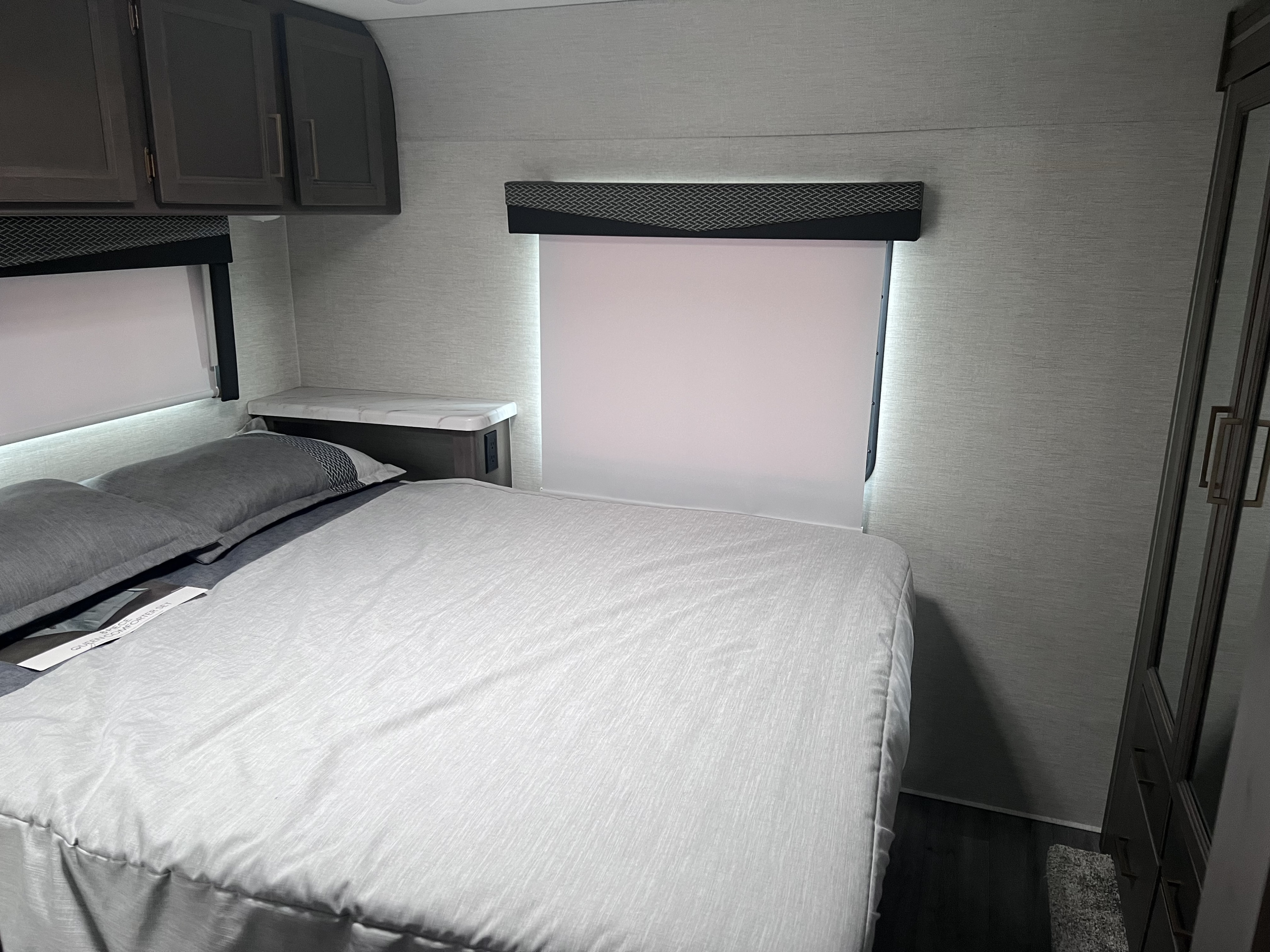 Comfortable RV rental bedroom with grey bedding, overhead cabinets, and windows with blinds. A modern, compact travel space.. Thor Four Winds 2024