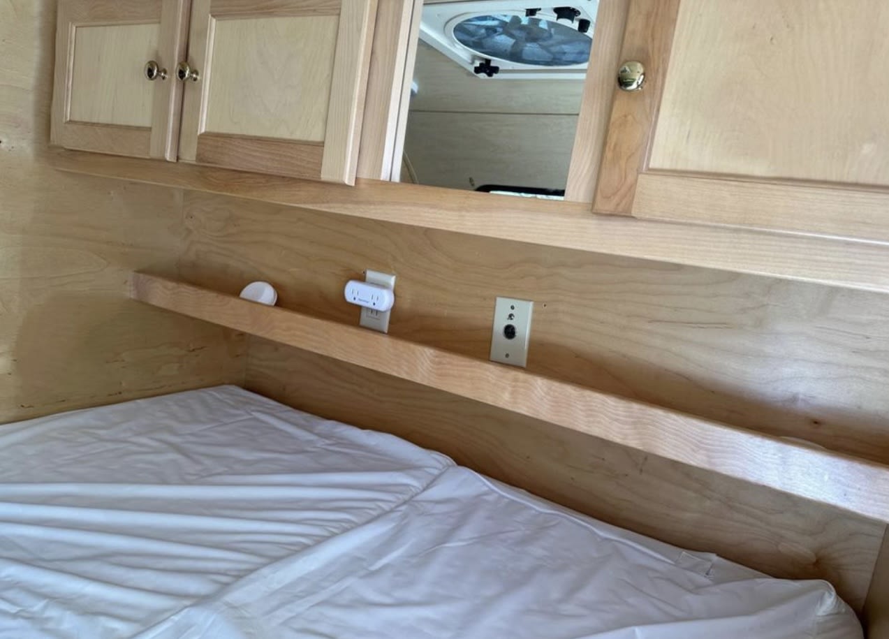 Cozy RV rental bedroom: wooden interior, white bed, shelf with power outlets & switch, overhead cabinets, and a roof vent.. Little Guy Silver Shadow 2011