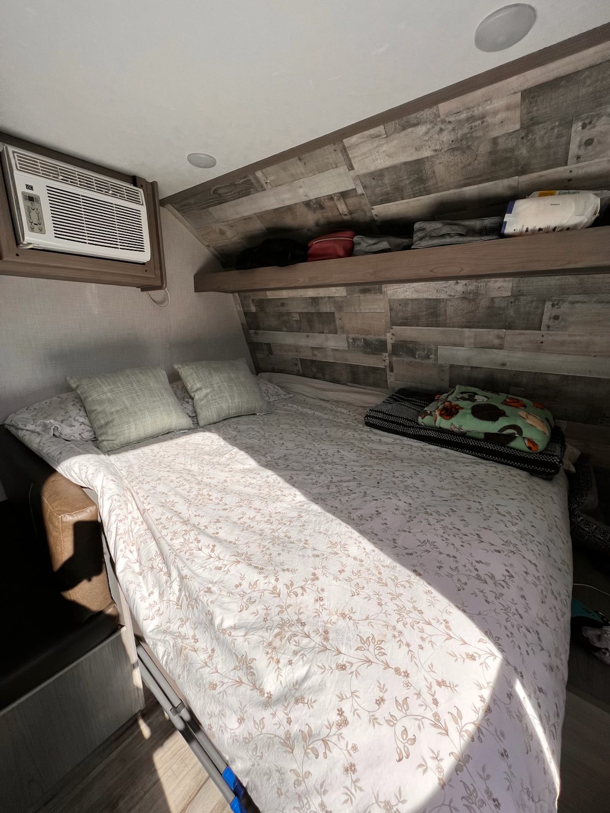 Cozy RV rental bedroom: sunlit bed with floral comforter, window AC, and wooden paneled walls & shelf.. Dutchmen Coleman Lantern LT 2023