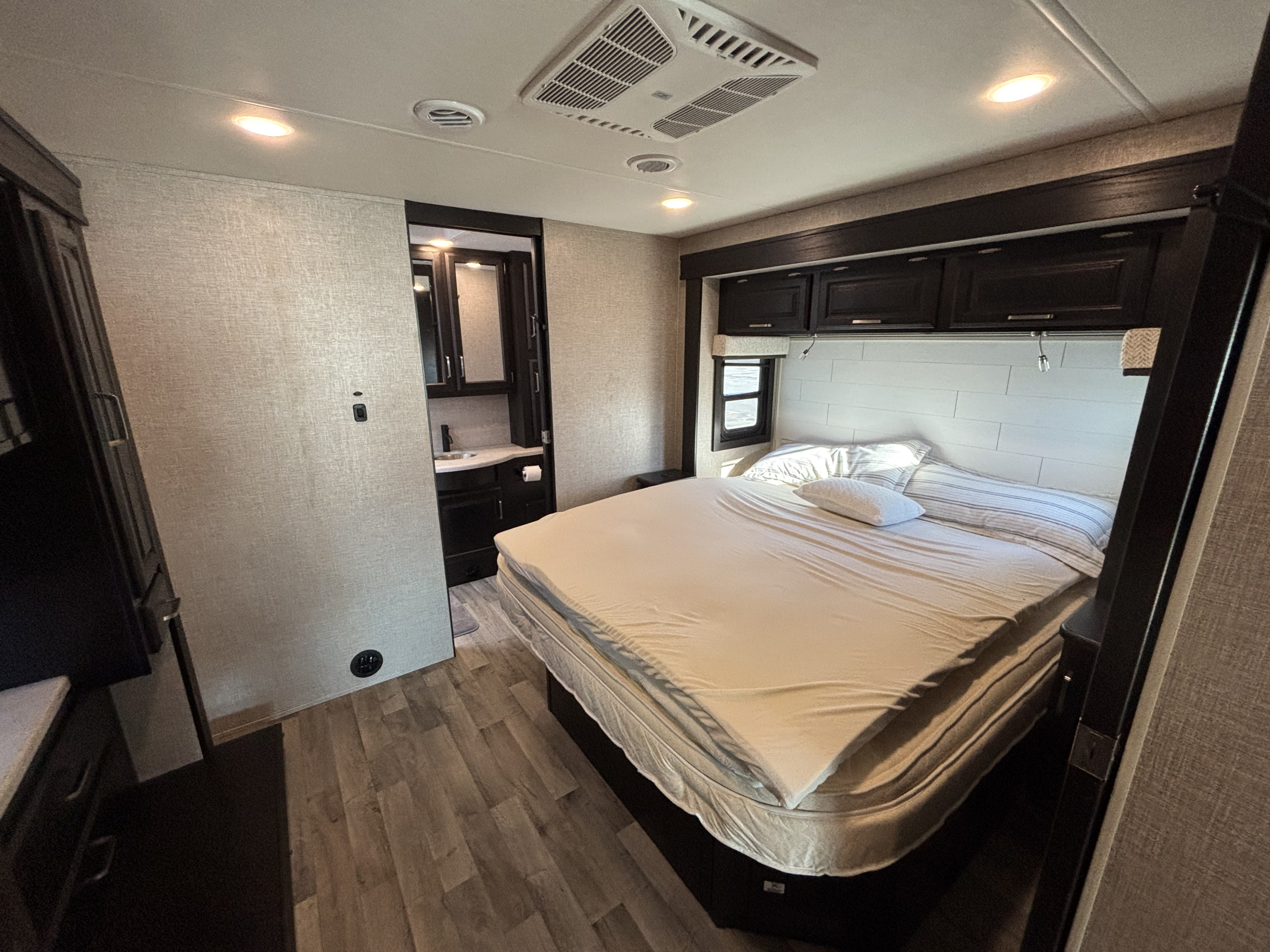 Well-lit RV rental bedroom with light bedding, dark overhead cabinets, and attached bath. Wood-look flooring and neutral walls.. Jayco Precept 2023