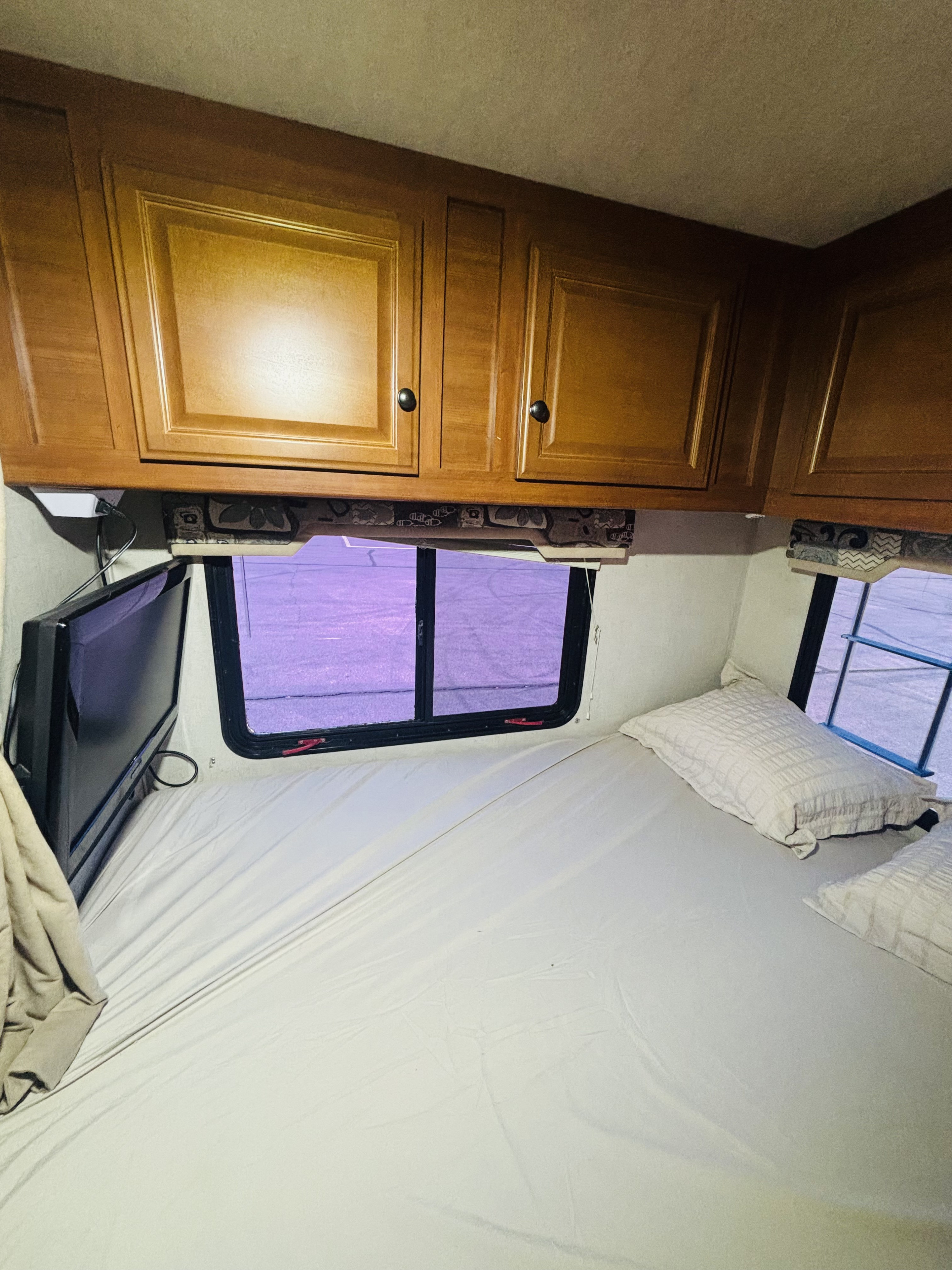 Inside an RV rental: a bedroom with a neatly made bed, overhead cabinets, a TV, and windows with a purple tint.. Jamboree Sport 2011