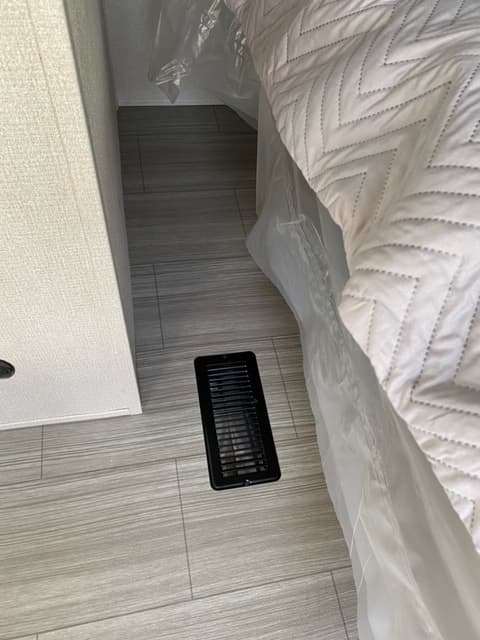 Inside an RV rental: light gray wood-look plank floor, black HVAC vent, and bed with white quilted comforter and protective plastic.. Keystone RV Hideout 2021