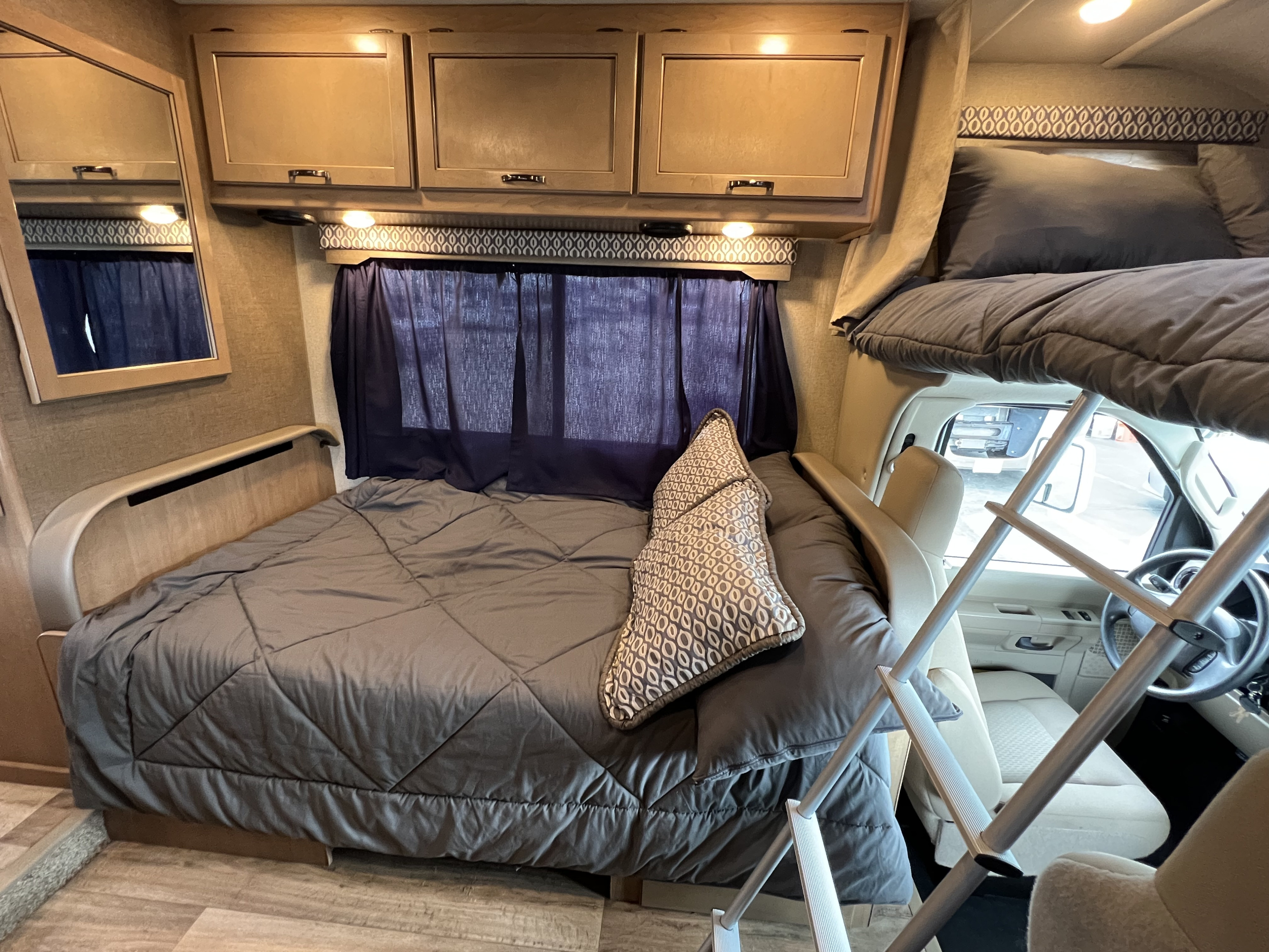 Inside an RV rental: a comfortable main bed, dark curtains, overhead storage, and a front bunk bed with a ladder to the driver's area.. THOR MOTOR COACH Freedom Elite 2019