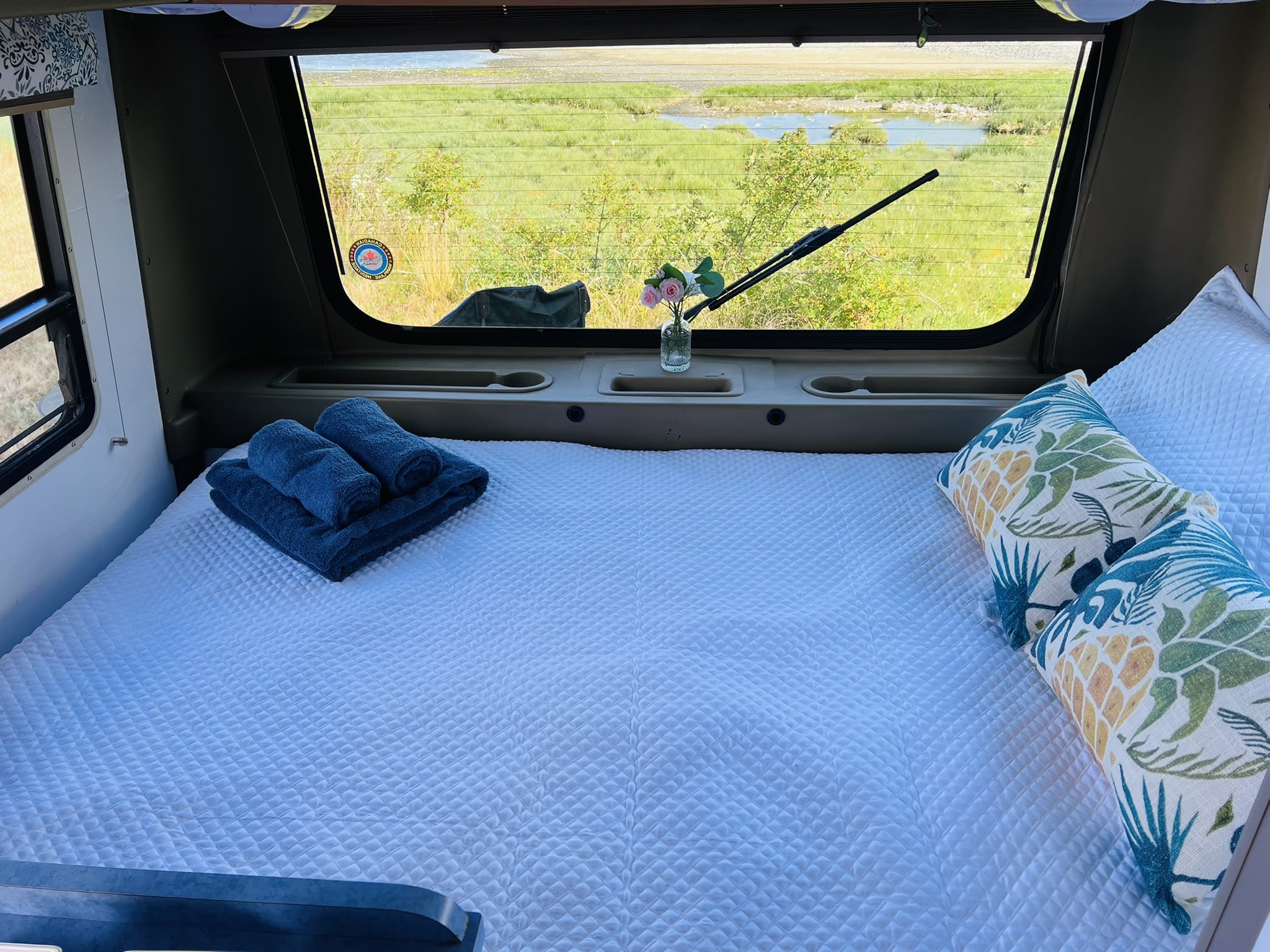 Inside an RV rental: A neatly made bed with white bedding, blue towels, and decorative pillows, offering scenic window views.. Volkswagen Westfalia 1995