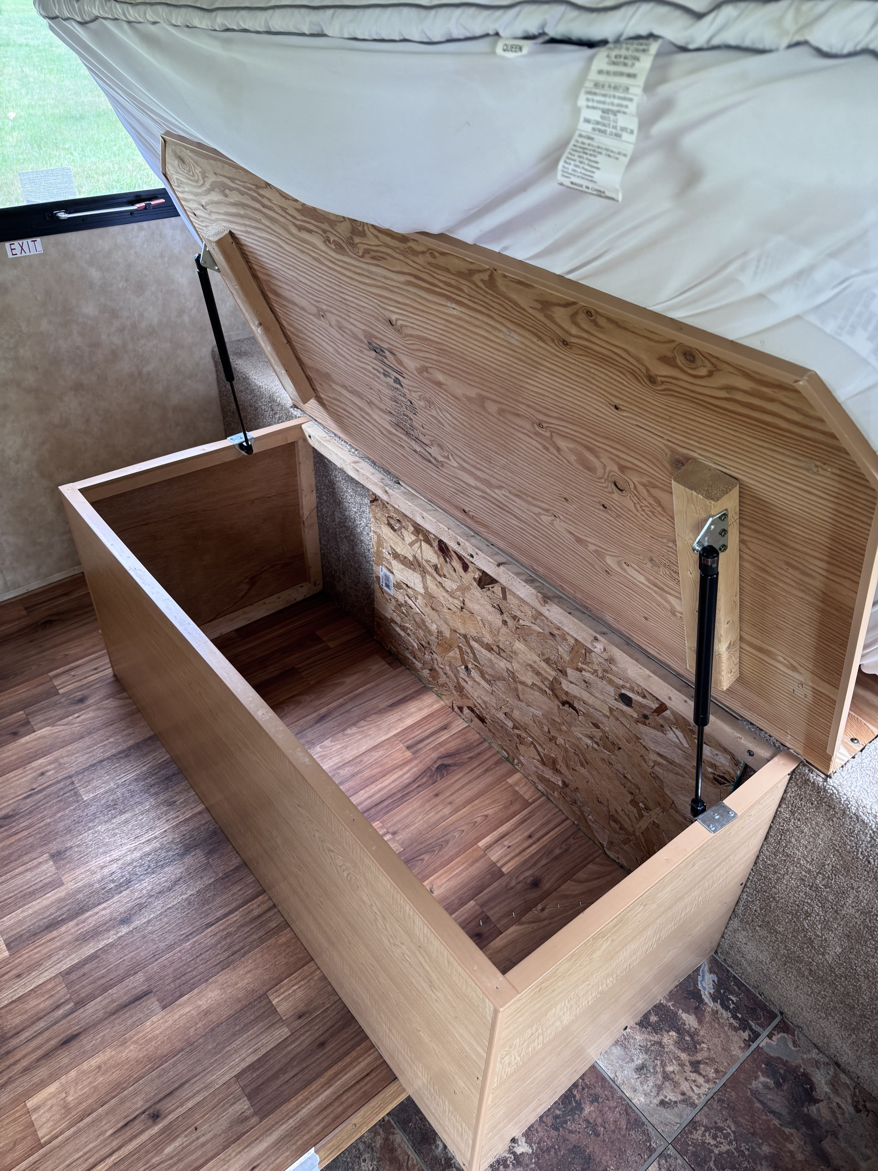 RV rental storage: Under-bed compartment with gas struts open, showing wood and OSB interior, queen bed above, exit sign.. kz Coyote 2010