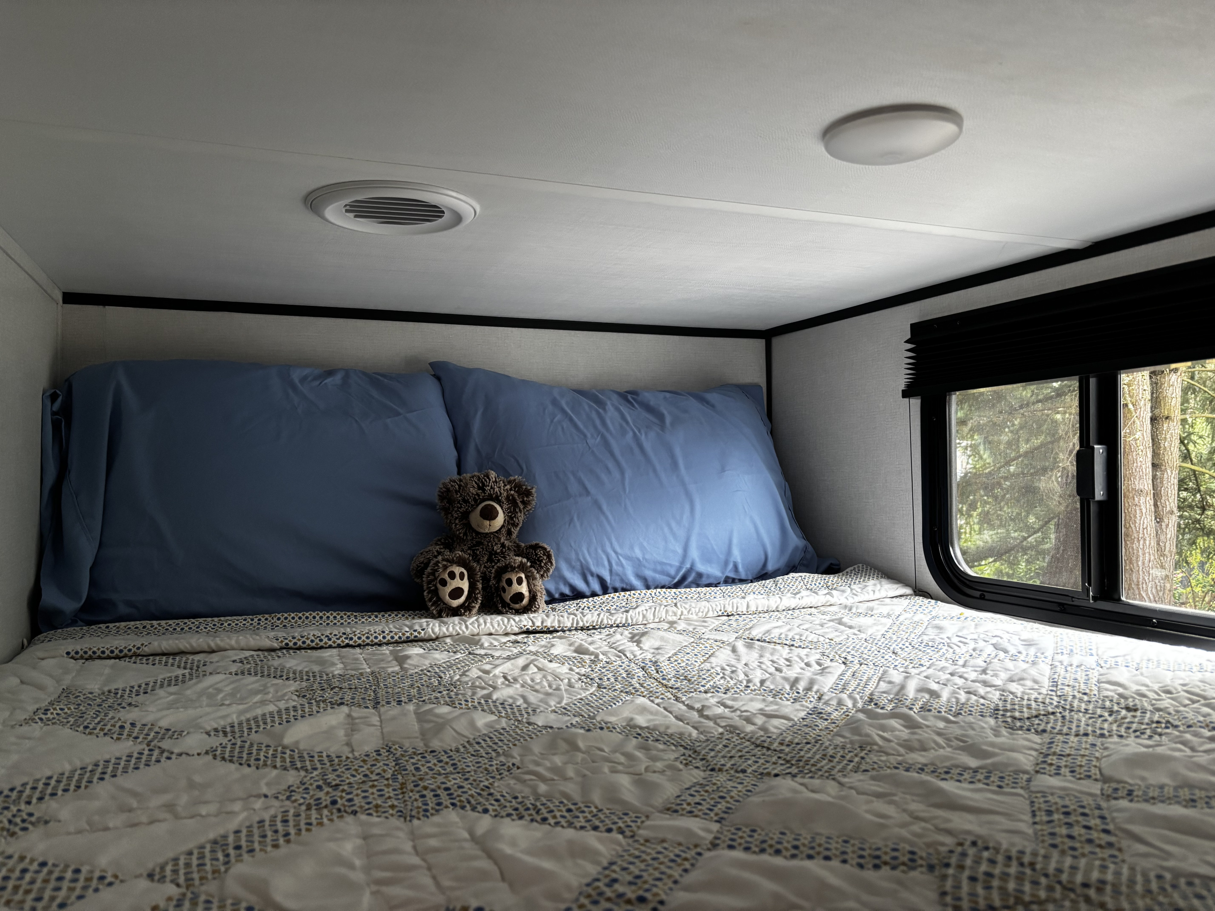 Cozy RV rental bed with blue pillows, patterned quilt, a teddy bear, and window view of trees.. Keystone Coleman Lite 22BX 2025