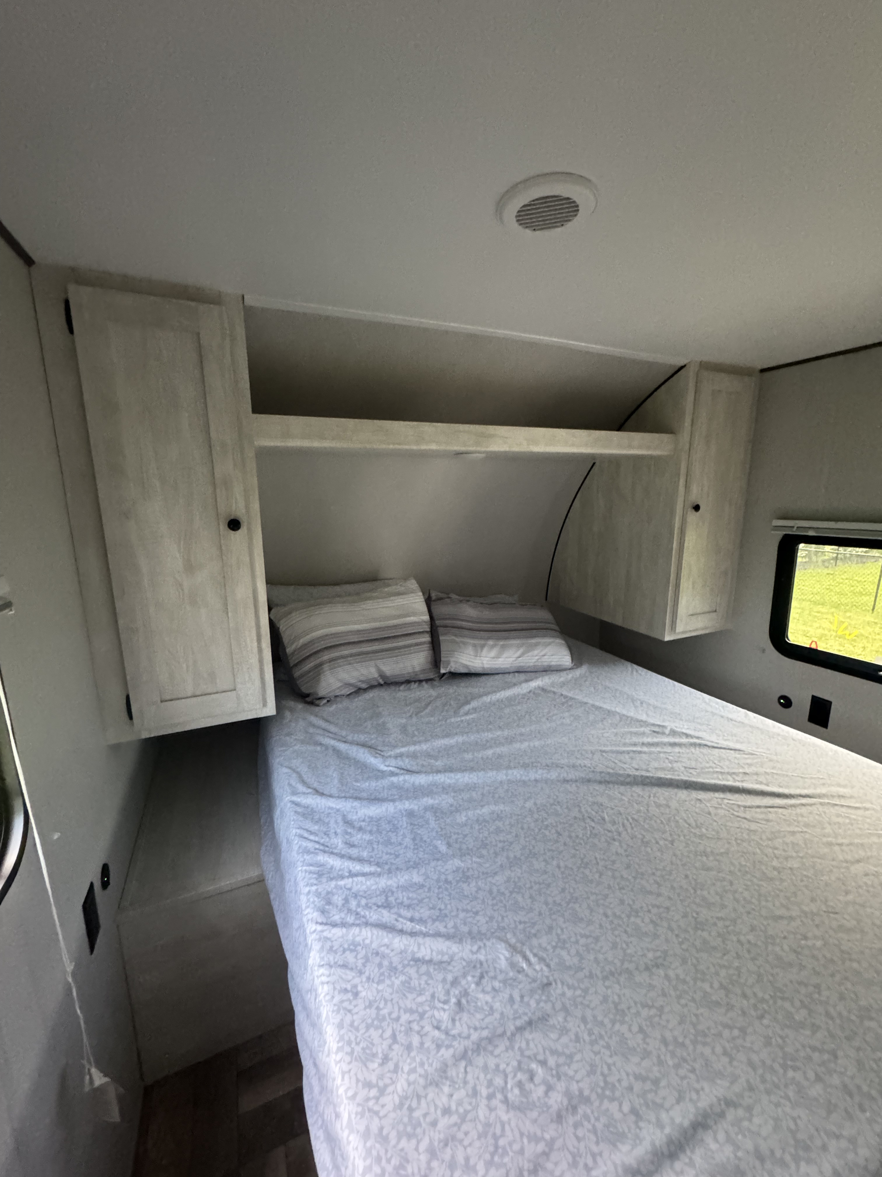 Inside an RV rental: a bright bedroom with a made bed, patterned sheets, striped pillows, and light wood overhead cabinets.. Forest River Shasta 2023