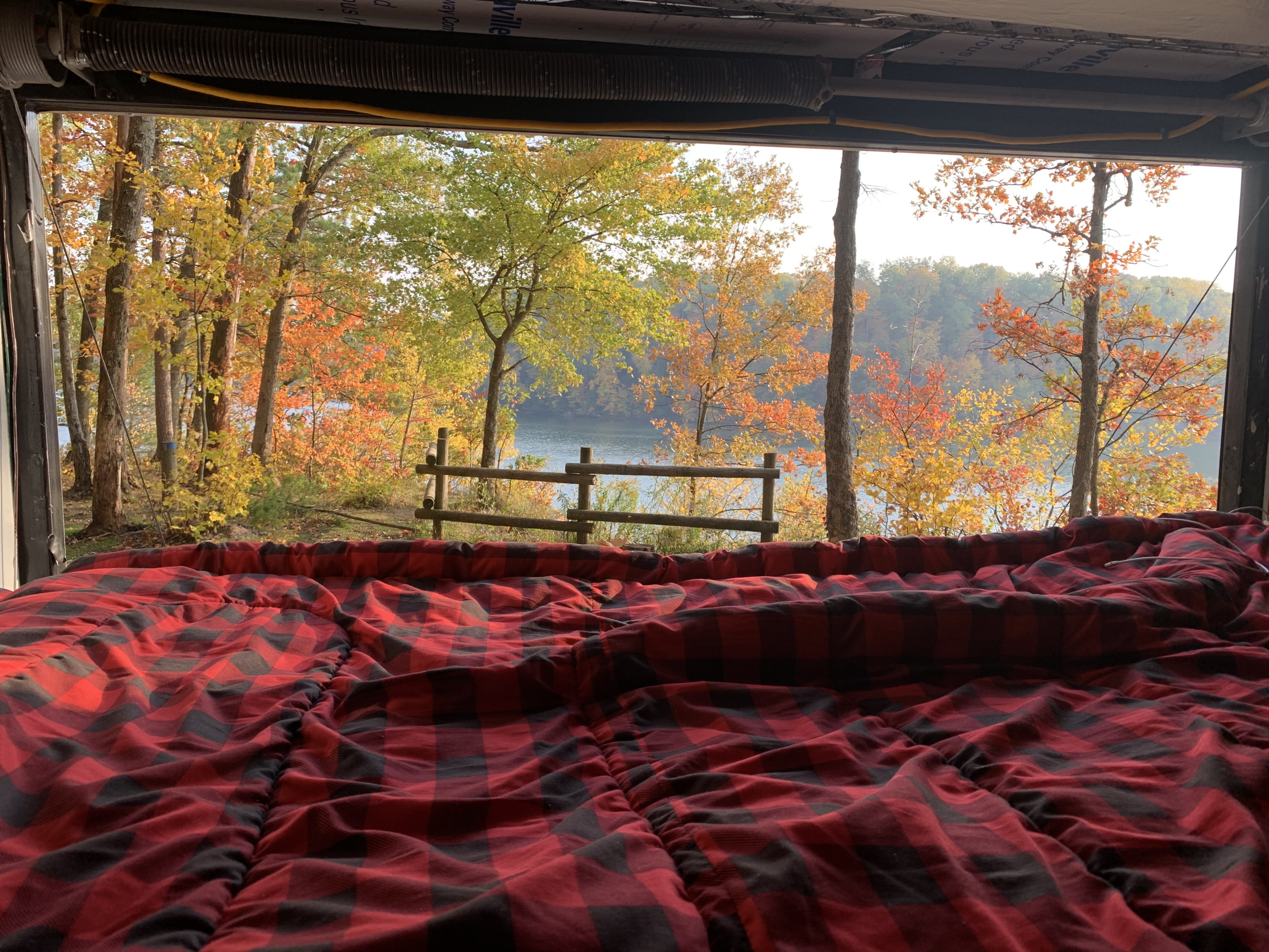 Cozy RV rental bed with a stunning autumn lake view. Red plaid comforter and vibrant fall trees create a perfect nature escape.. impact trailer itb714ta 2019