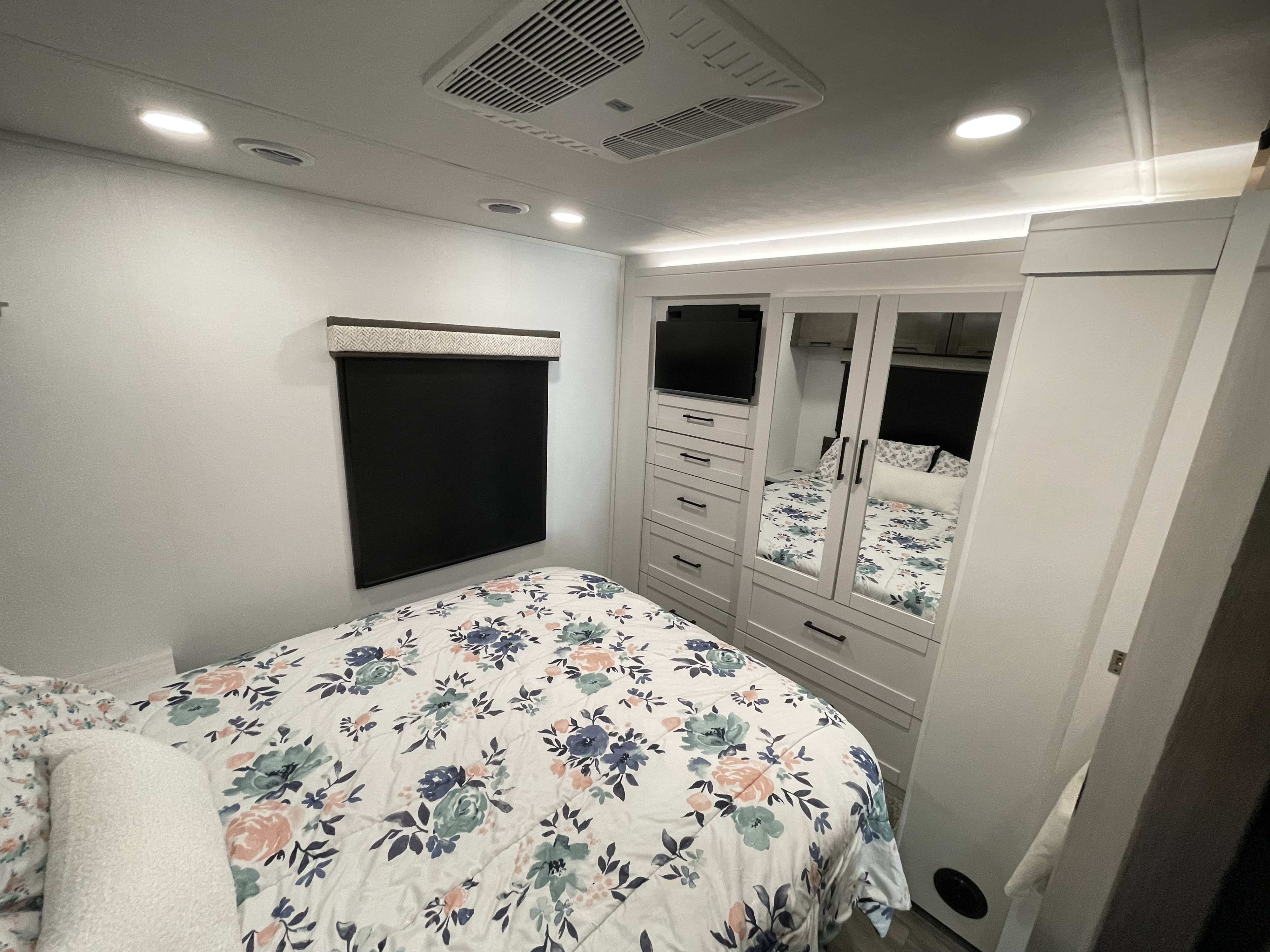 Cozy RV rental bedroom featuring a floral comforter, built-in storage, TV, window, and AC unit. Modern & functional.. Jayco Greyhawk 2026