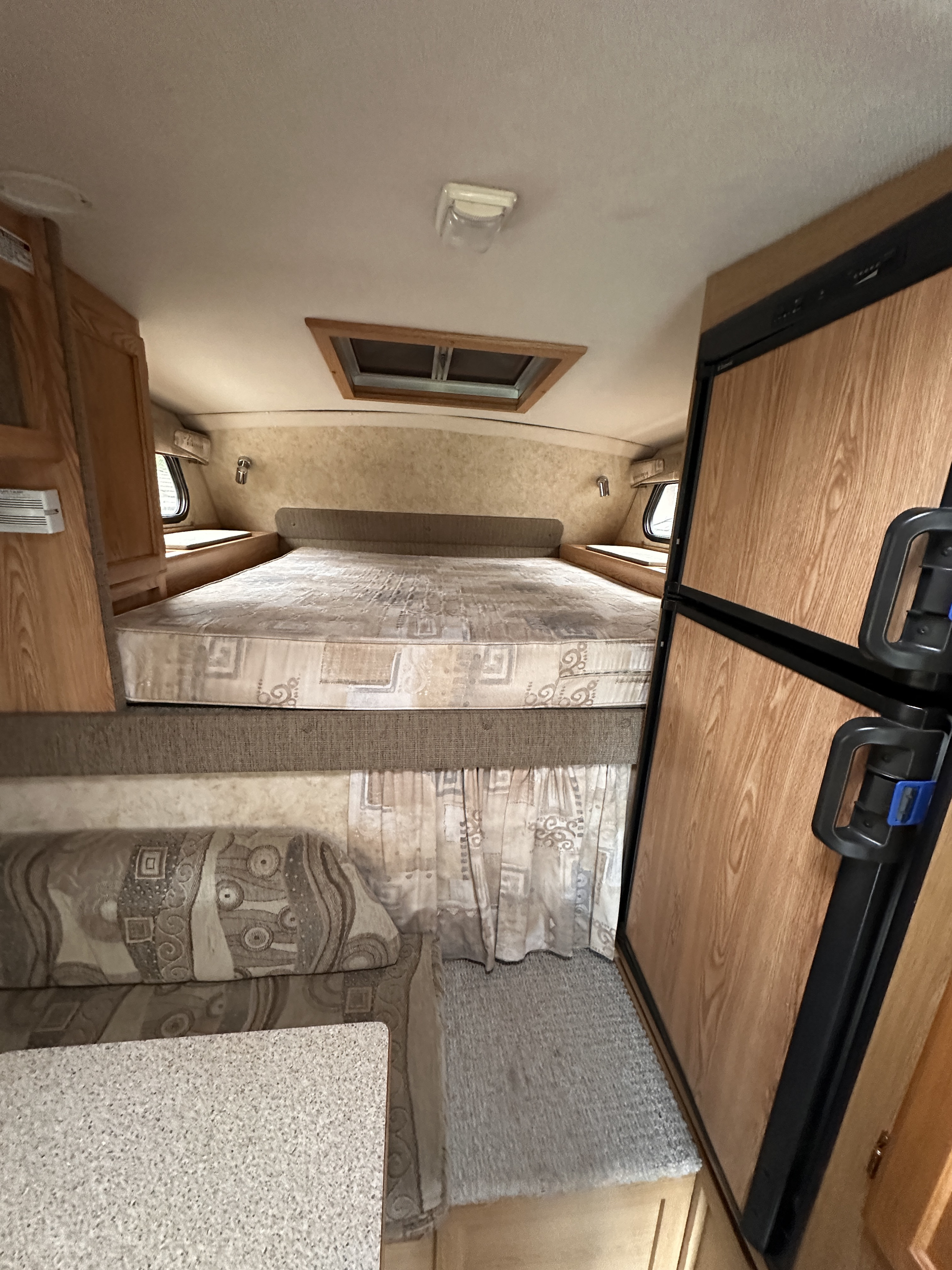 Cozy RV rental interior featuring a comfortable bed, compact refrigerator, and lounge seating with a table.. Bigfoot 1500series 2006