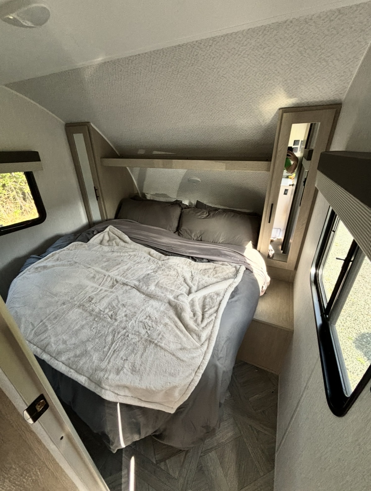Cozy RV rental bedroom featuring a comfy bed with grey bedding, mirrored storage, and windows showing outdoor scenery.. Forest River Wildwood X-Lite 2024