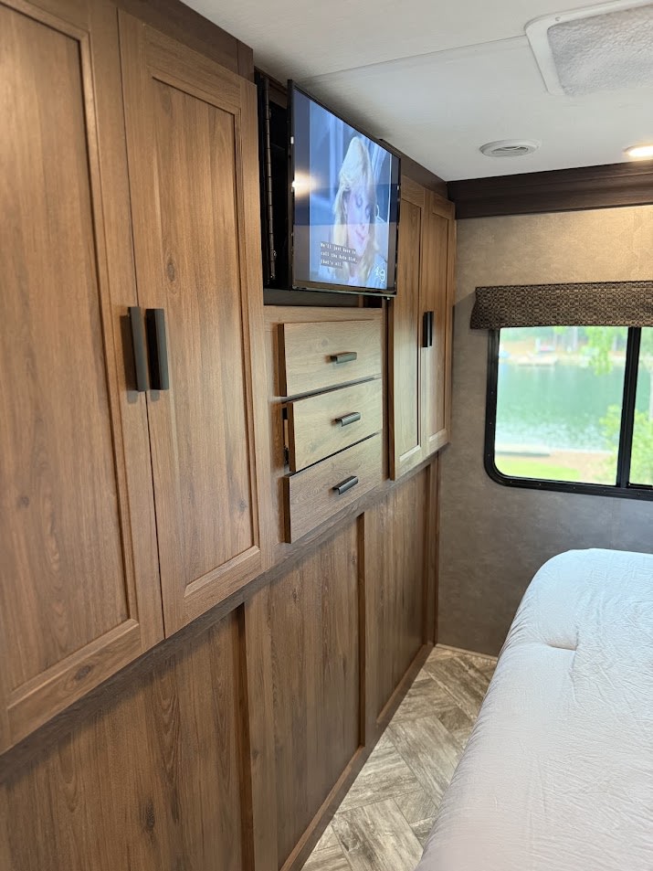RV rental bedroom: wood cabinetry, wall-mounted TV, open drawer storage, and a picturesque lake view.. Forest River Sunseeker Classic 2022
