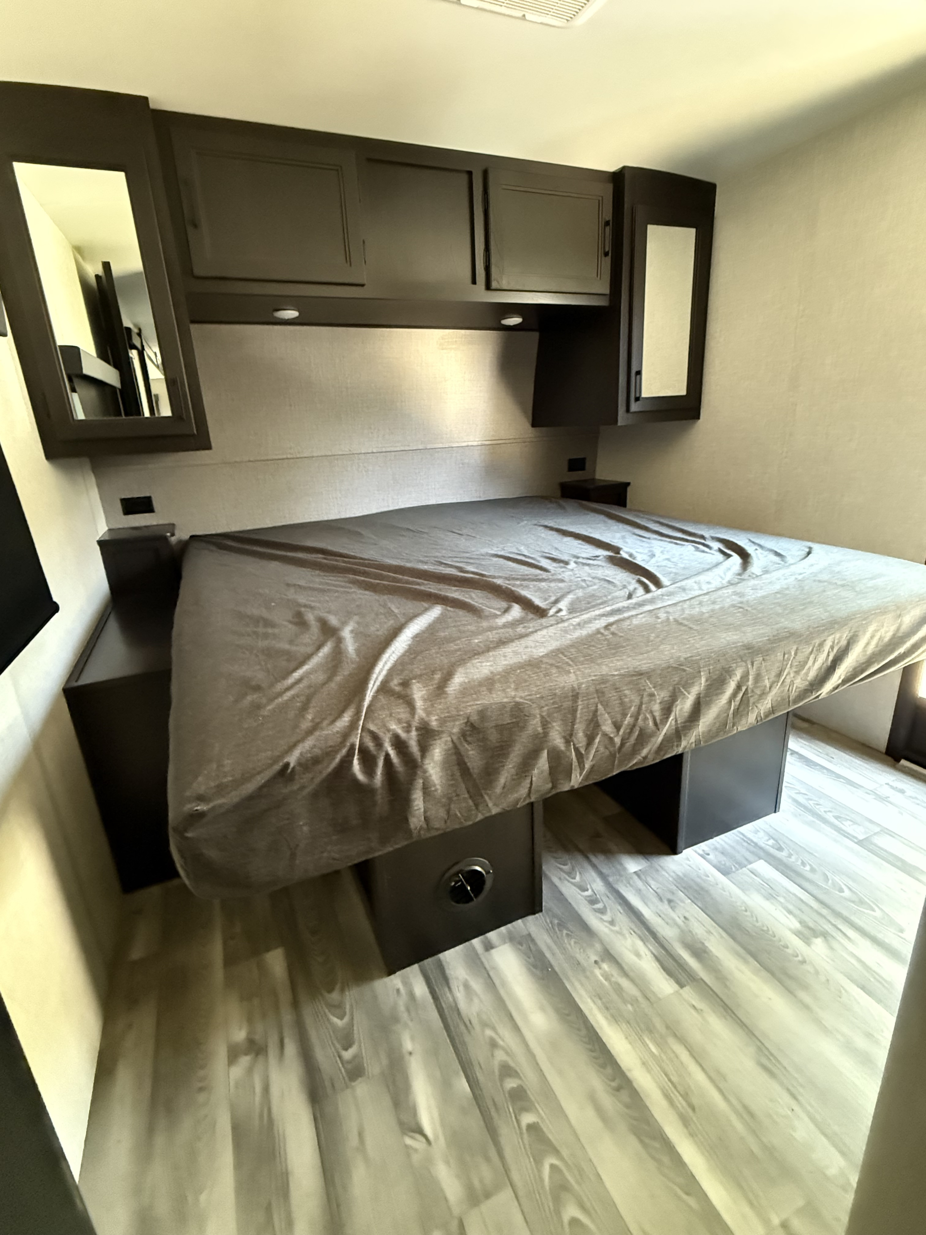 This RV rental bedroom features a spacious bed with a grey sheet, dark overhead cabinets, and built-in nightstands. Light wood-look flooring is visible.. Starcraft 296bhs 2023