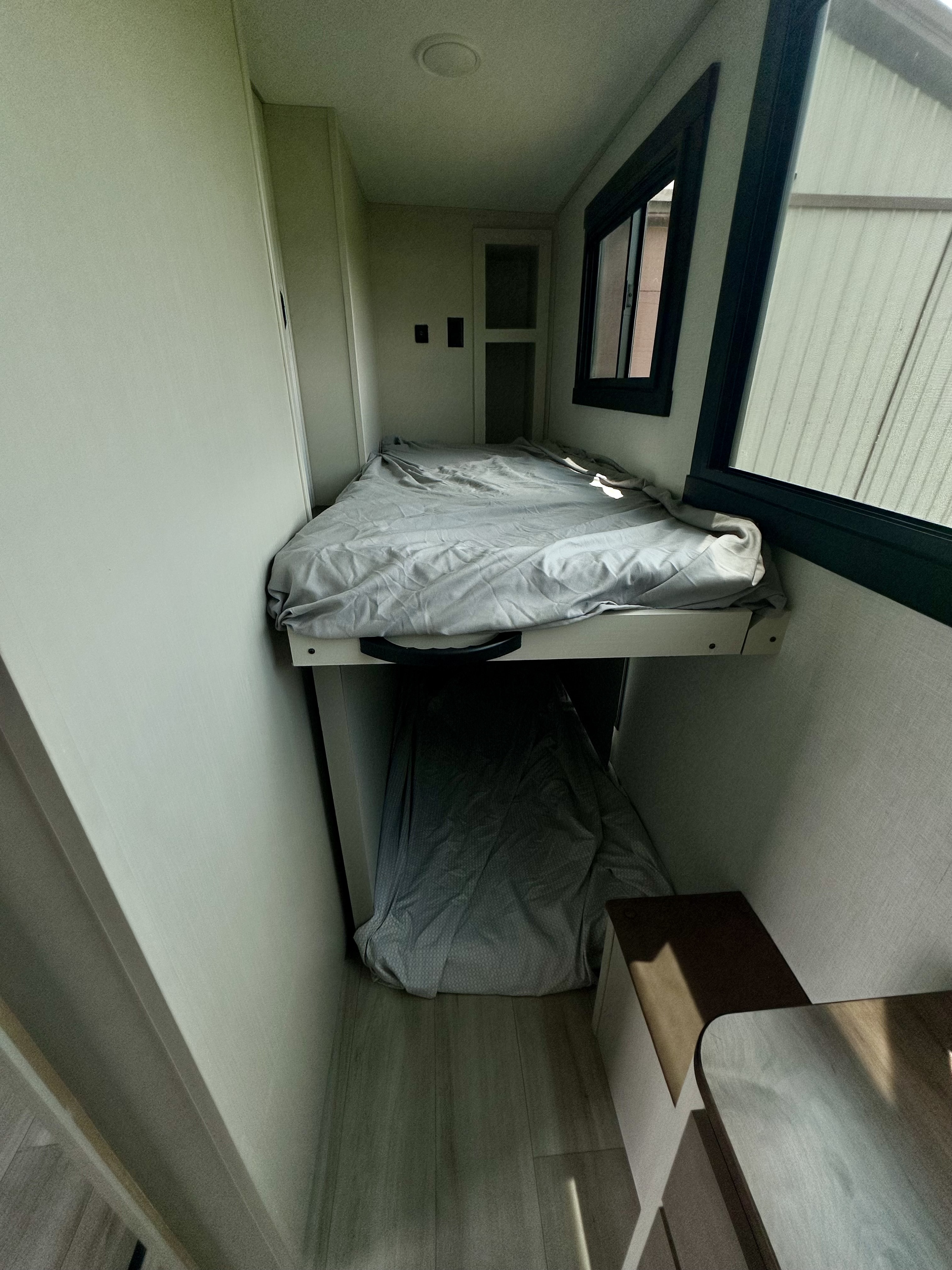 Compact bunk beds in an RV rental sleeping area. Upper and lower bunks with grey bedding, plus a window on the right.. Forest River Heritage Glen 2025