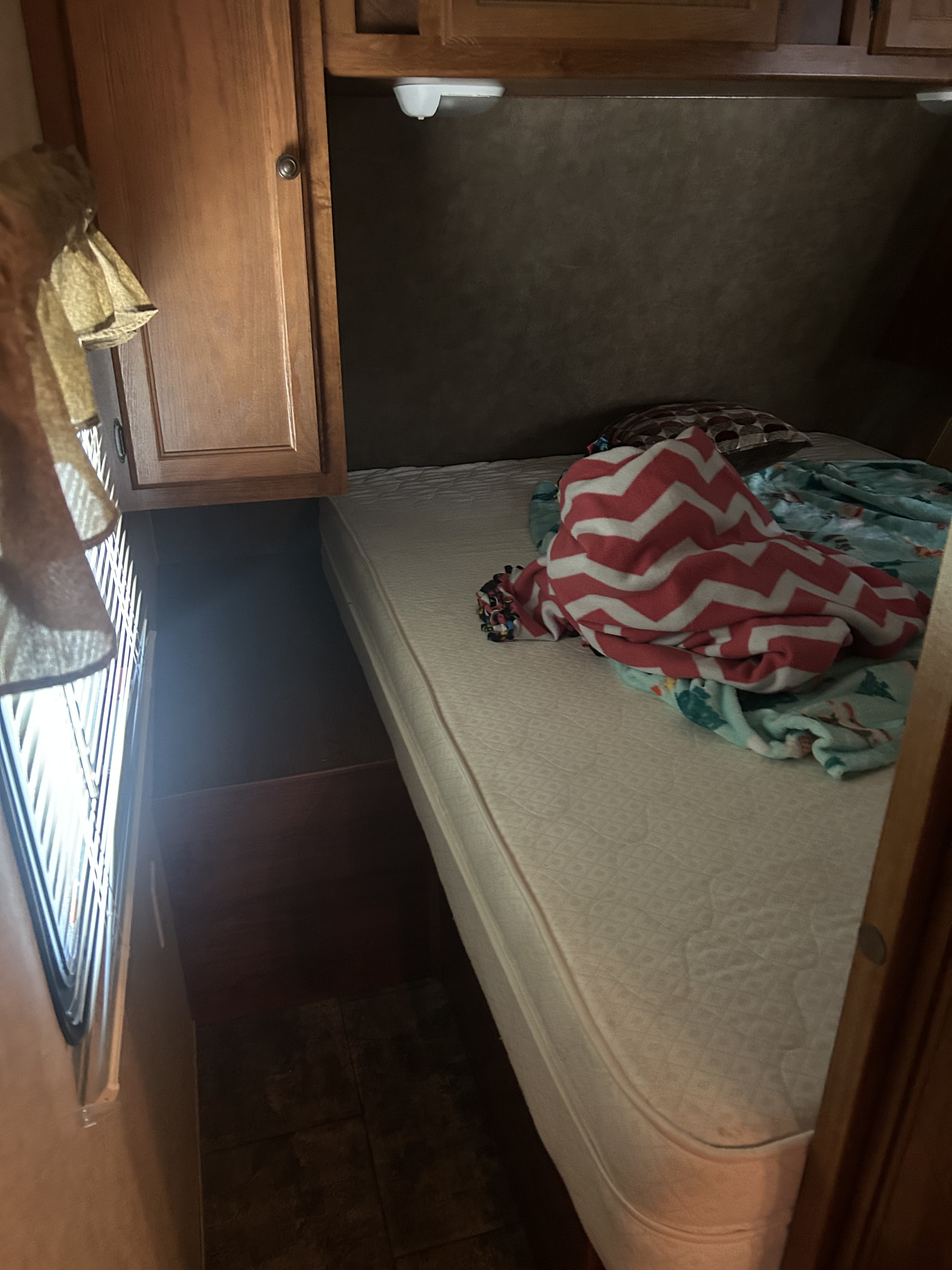 Compact bedroom in an RV rental, featuring a mattress with cozy blankets, wooden cabinets, and window light.. Heartland Trail Runner 2013