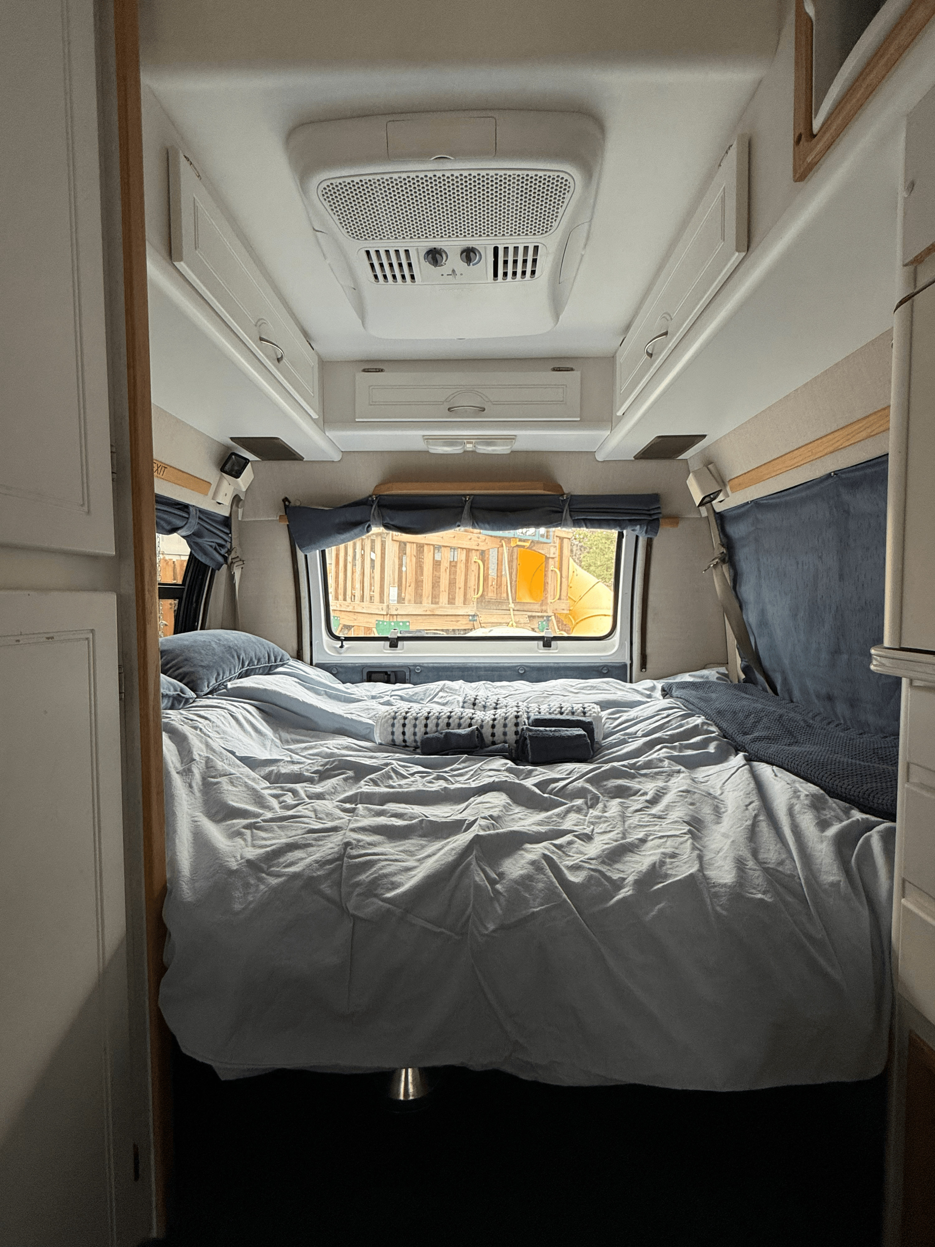 An RV rental's cozy sleeping area with a made bed, overhead AC, storage, and a playground outside the rear window.. Pleasure Way Excel 2001